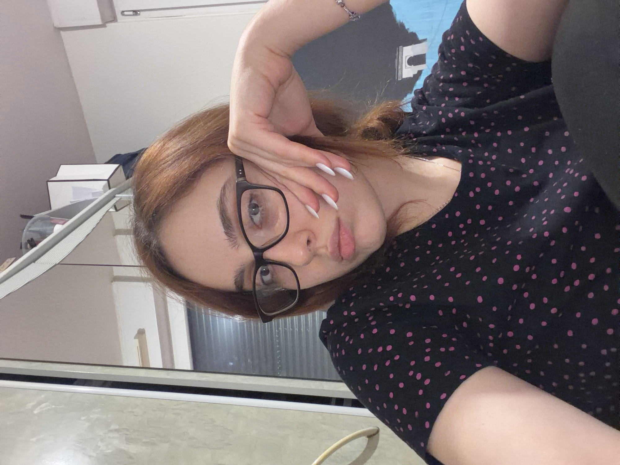 A person with light brown hair is wearing glasses and a black shirt with pink polka dots. They are resting their head on their hand, which has long white nails. The background includes a white box and a window. The location is not discernible from the provided image.