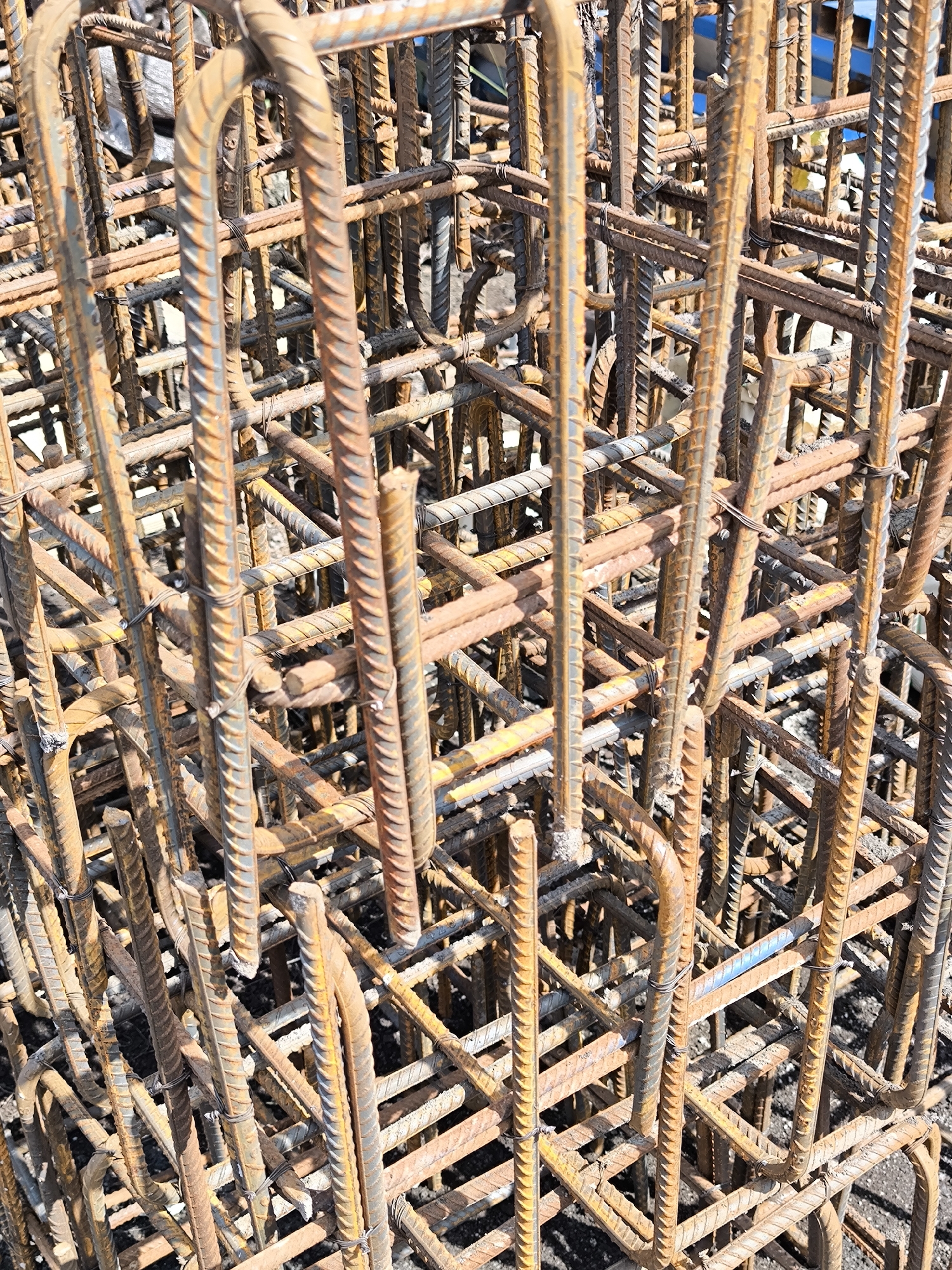 The image shows a stack of prefabricated reinforcement cages made of ribbed steel rebar. The cages are rectangular prisms with vertical bars bent into U-shapes at the top. Horizontal rebar is tied to the vertical bars with thin wire. The rebar has a rusty, oxidized surface. The location appears to be a construction site, likely in Unknown, Unknown. No people are visible.