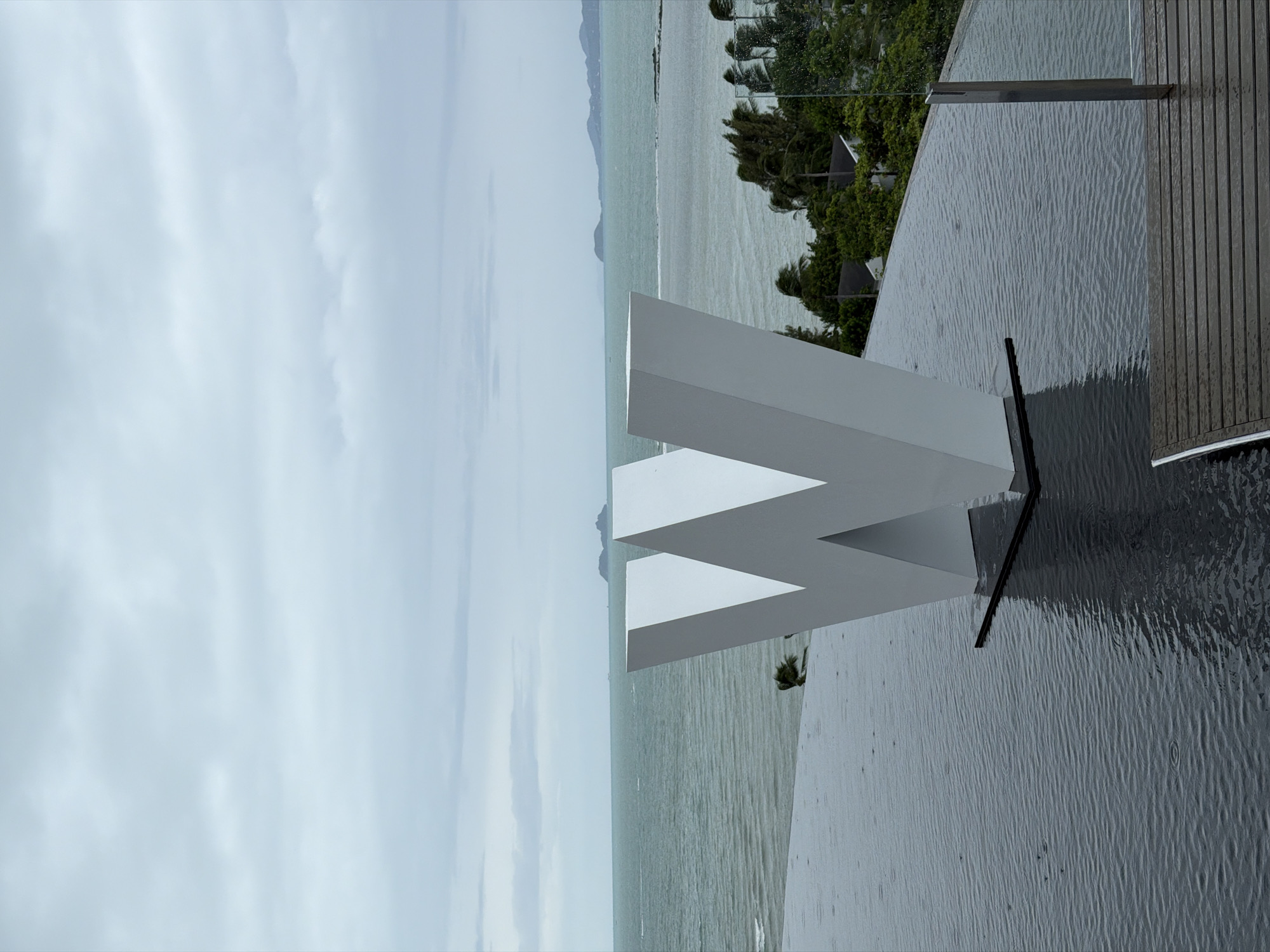 The image depicts a large, white, three-dimensional letter "W" sculpture positioned in the foreground of an infinity pool. The pool's surface is dark and rippled, reflecting the overcast sky and the sculpture. Beyond the pool, a vast expanse of the ocean stretches towards the horizon, where several islands and a hazy coastline are visible. Trees and some buildings are situated on a lush green hill to the right of the frame. The sky is a uniform pale gray, indicating cloudy weather, and the ocean is a muted greenish-blue. The overall impression is one of a tranquil, somewhat somber, tropical vista. This scene is consistent with the views from luxury resorts in the Bophut or Maenam areas of Ko Samui District, Thailand, known for their oceanfront infinity pools and dramatic landscapes.
