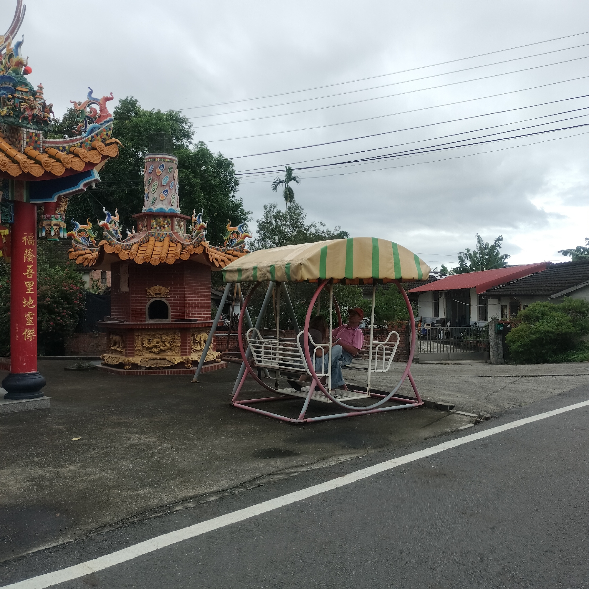 The image depicts a street scene in Zhushan Township, Taiwan, on an overcast day. In the foreground, a paved road with a white line marking runs horizontally across the frame. To the left, a paved courtyard area contains a colorful, ornately decorated temple structure with a red pillar bearing Chinese characters. Adjacent to the temple is a brick structure with a curved roof and an arched opening, also adorned with intricate carvings. In the middle ground, a swing set with a striped canopy is present, with two individuals seated on it. One person, wearing a pink shirt and a red cap, is looking at a mobile phone. Behind the swing set, a residential house with a red roof and a dark tiled roof is visible, partially obscured by dense green foliage. Overhead, multiple power lines are strung across the sky, which is filled with grey clouds. The environment appears to be a semi-rural or suburban area.