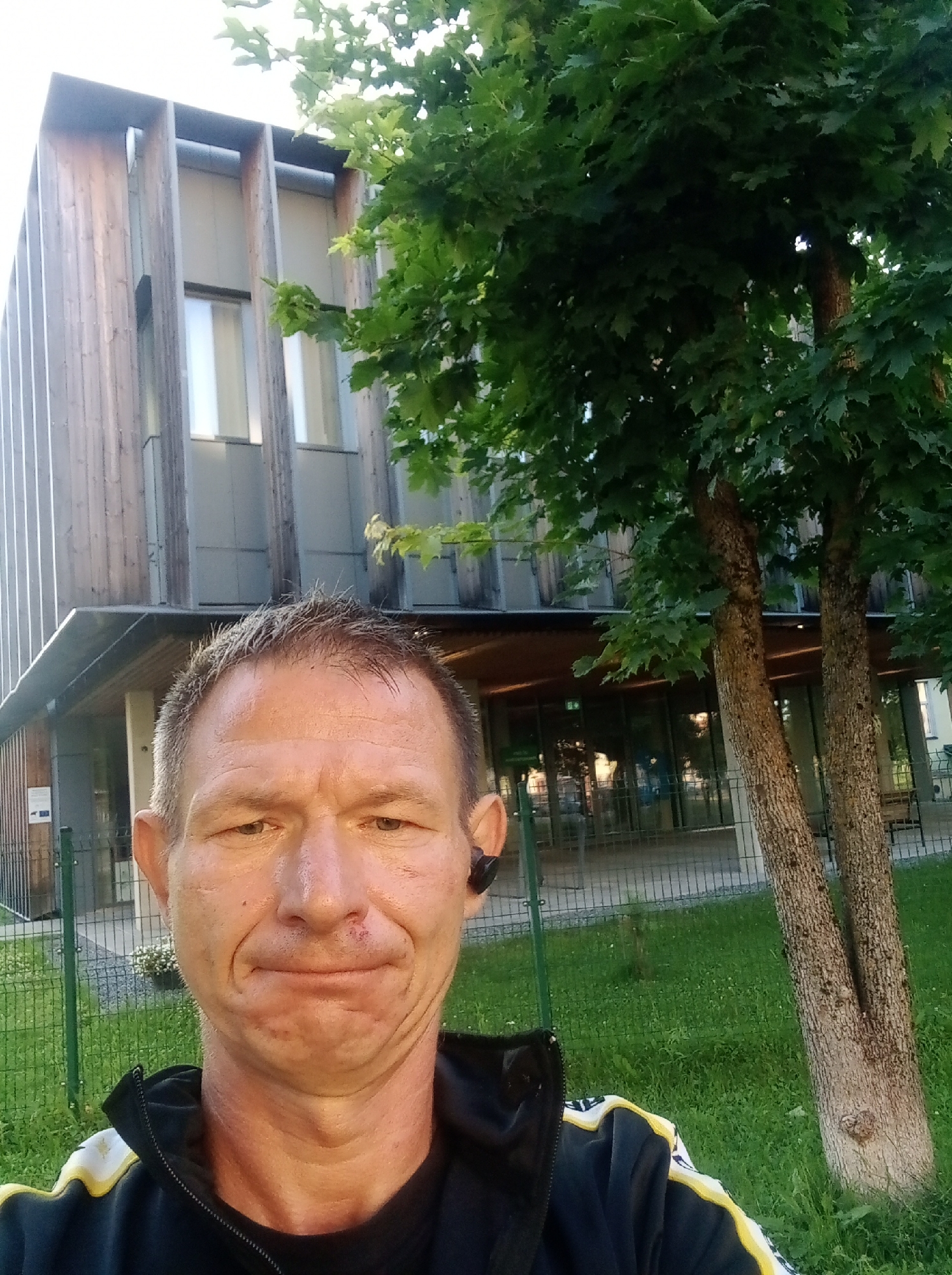 A man is standing in front of a modern building with wooden accents and large windows. The building appears to be a public institution, possibly a school or community center. There is a tree with green leaves in the foreground, and a fence surrounds the property. The man is wearing a black jacket with yellow and white accents. He is looking at the camera with a neutral expression. The image was taken in Tapa, Estonia.