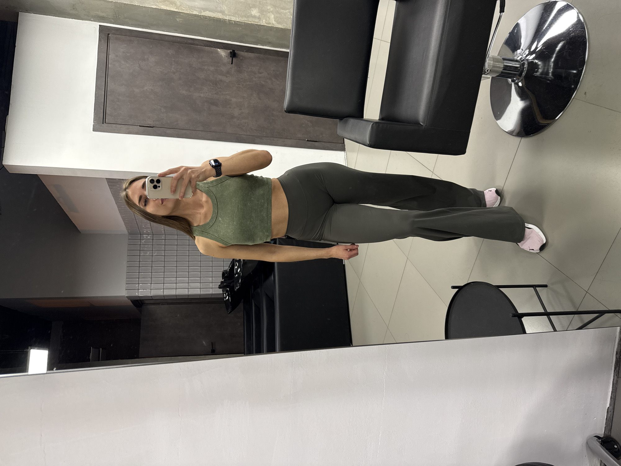 A woman takes a mirror selfie in a modern, industrial-style indoor setting. She stands, partially obscured by the phone she holds to her face, wearing an olive green ribbed crop top, dark gray-green flared athletic pants, and light pink and white sneakers. A black digital smartwatch is visible on her left wrist. Her gaze is directed towards the phone, and her expression appears neutral.
The background features clean lines and neutral tones, including a large dark gray-brown door or panel set into a white wall, a raw concrete ceiling detail, and a floor laid with large gray tiles. On the right, a black salon-style chair with a reflective silver hydraulic base is prominently positioned. A small, black circular table with metal legs is partially visible in the bottom right corner. The overall atmosphere suggests a contemporary commercial space such as a salon, gym, or studio, possibly located in Saint Petersburg, Russia. The image documents a casual self-portrait, showcasing an outfit or a moment in the space.