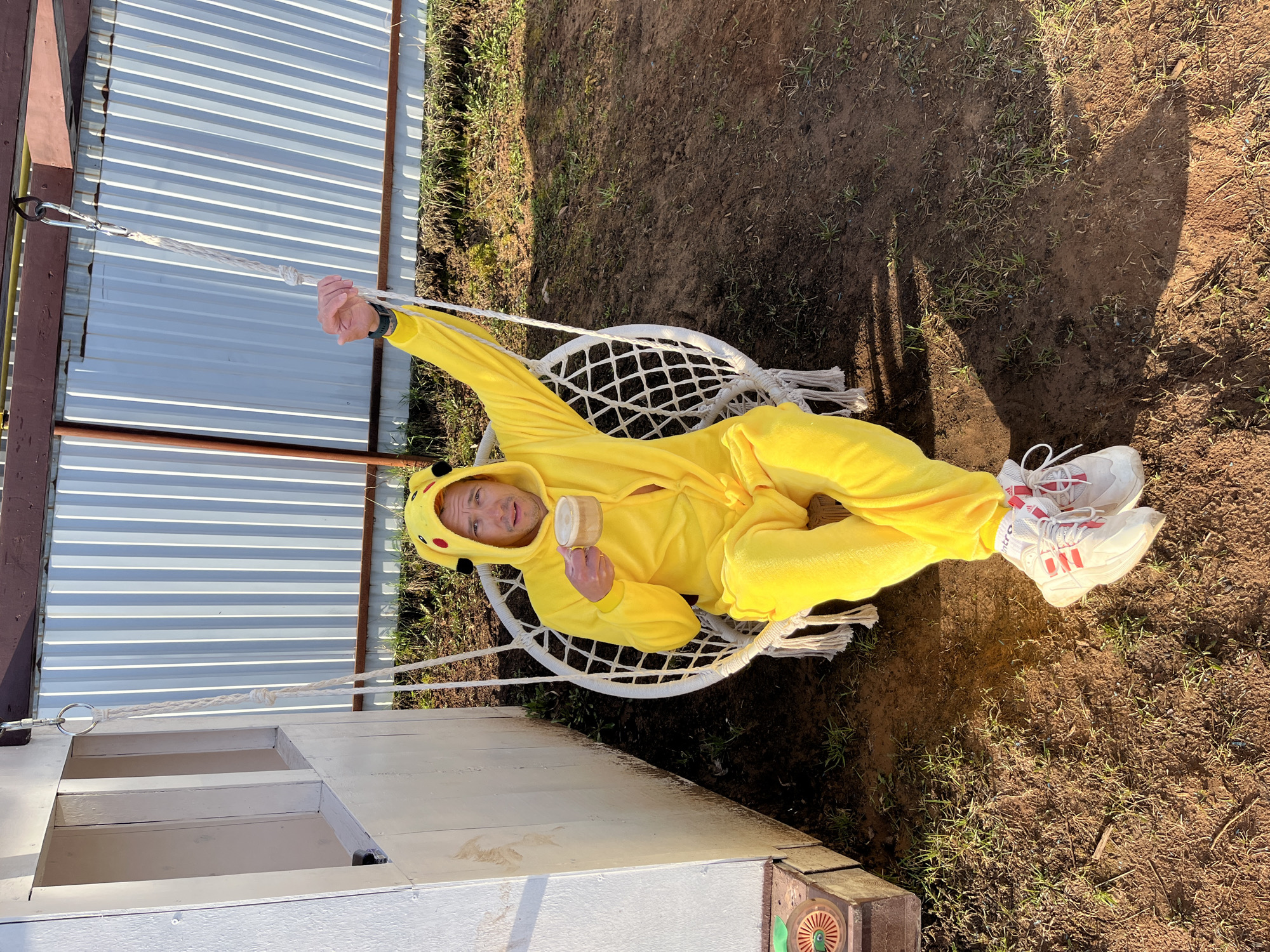 A man wearing a Pikachu costume is sitting in a white macrame swing attached to a wooden structure outdoors. He is holding a small wooden object in his left hand. The location appears to be a yard in Chaikovsky, Russia, with a corrugated metal fence and grass. The scene is lit by sunlight.