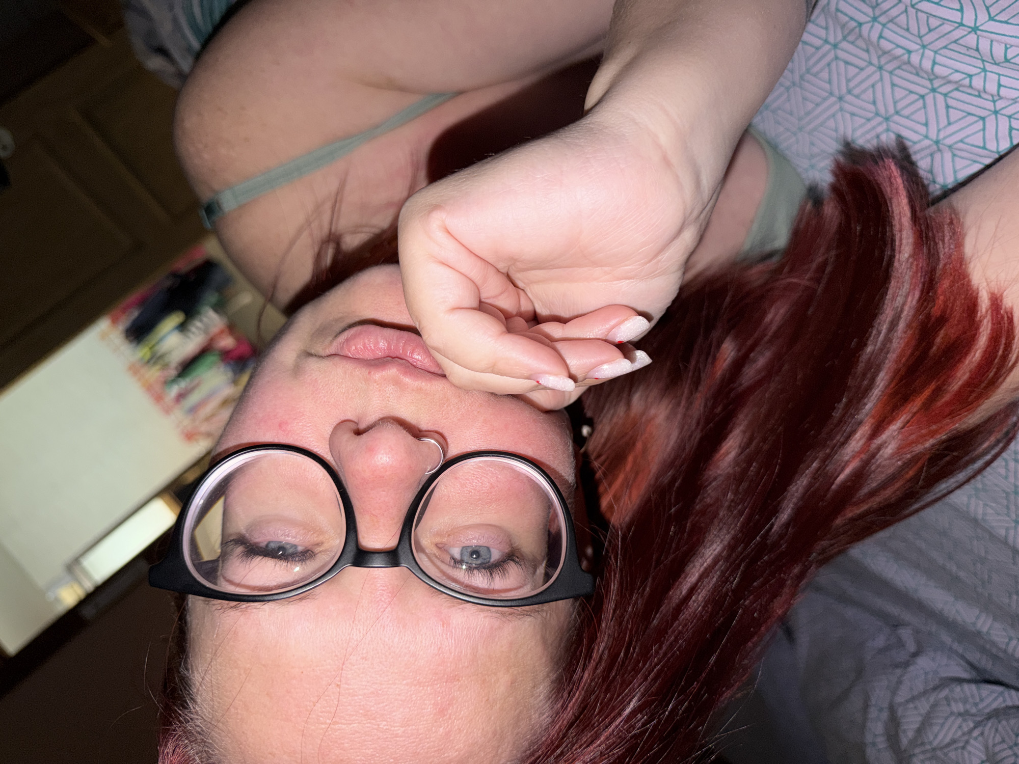 A close-up, upside-down image shows a person with red hair, wearing black framed glasses, and lying on a bed with patterned sheets. The person's hand is near their face. A partially visible object is on the left of the image, near a wooden door. The city of Chaikovsky, Russia, is not discernible from the image.