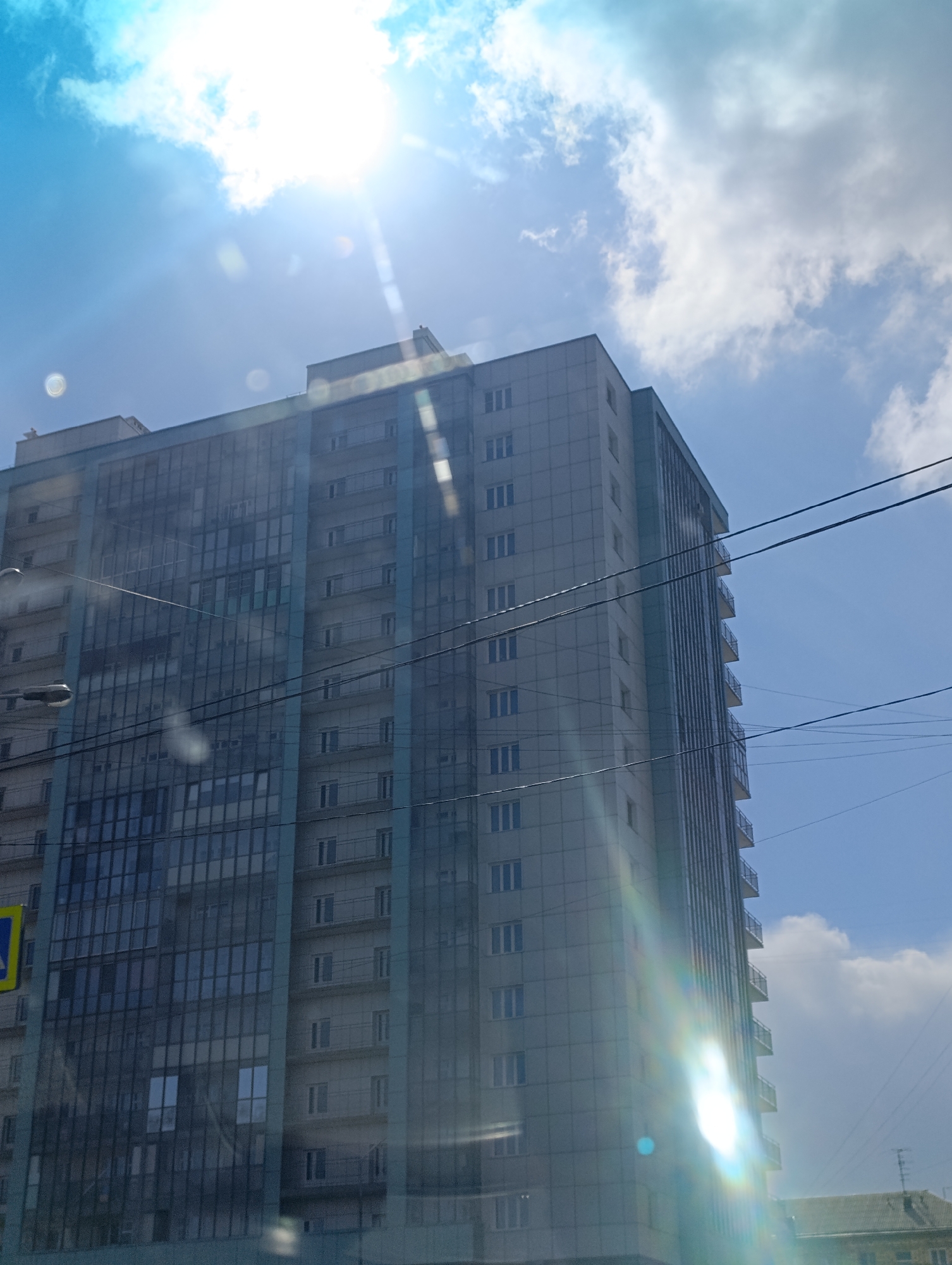 The image shows a tall apartment building in Krasnoyarsk, Russia under a bright, slightly cloudy sky with the sun shining through. The building appears to be made of concrete and glass, with rows of windows and small balconies. Power lines are visible in the foreground. A street sign is partially visible on the left. Other buildings are partially visible at the bottom of the image.