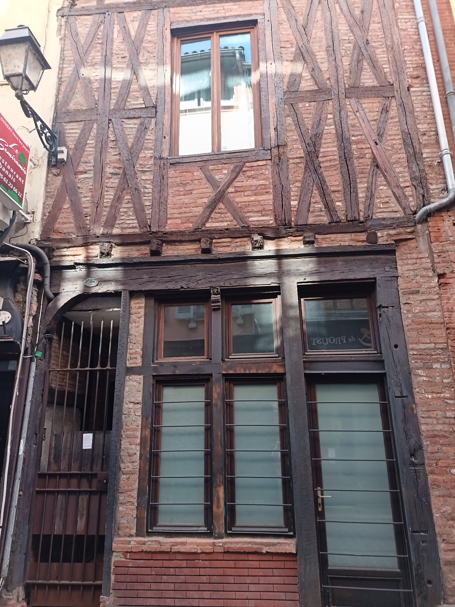This image captures the facade of a multi-story, half-timbered building, showcasing traditional architectural elements typical of historic European cities like Toulouse. The structure features a dark wooden framework with intricate, angular bracing patterns set against a rustic red brick infill.
The upper floor prominently displays large wooden beams forming X-shaped and vertical/horizontal configurations. A single window with a light wooden frame reflects the bright sky.
The ground floor features a more robust wooden beam structure. On the far left, a traditional black street lamp with an ornate bracket extends from the wall, next to a partially visible red commercial sign. This sign reads "OUVERT tous les jours", "Semiramis", "RESTAURANT Libanais", and a phone number "05 61 23 66 11". Below the lamp, an arched opening is secured by a dark, possibly rusty, metal grate or gate. Above this arch, a circular house number "25" is affixed. To the right, several ground-floor windows are visible, characterized by dark frames and internal blinds or horizontal security bars. One window distinctly displays the reversed text "de Proust" along with a faint rocking horse image, suggesting a shop or business.
A grey drainpipe runs vertically down the right side of the building. The overall scene suggests a static, daytime view with clear weather, emphasizing the aged character and detailed construction of the building. No discernible activity or people are present.