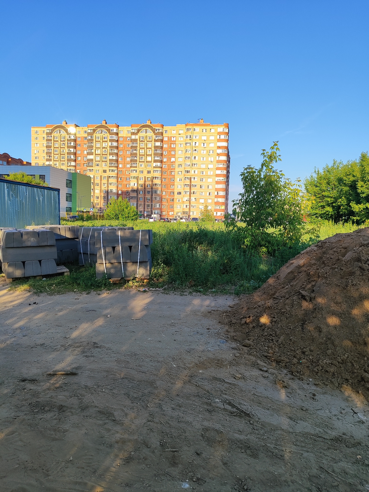 The scene depicts an outdoor urban landscape situated in Shchyolkovo, Russia, under a clear, bright blue sky.
In the background, a large, multi-story residential apartment building with an orange and light beige facade dominates, featuring numerous windows, some with enclosed and open balconies, and arched architectural details on its upper levels. Direct sunlight causes reflections on several windows, particularly on the upper-left side of the building. To the immediate left of this main structure, a smaller building with white and green sections is partially visible, alongside a segment of a reddish-brown roof further to the left.
The midground is characterized by an area of tall, vibrant green grass, interspersed with various shrubs and small trees. A yellow triangular road sign with a black pedestrian symbol is partially obscured within this vegetation. On the left side of the midground, multiple stacks of dark grey pre-cast concrete curbstones or paving blocks are neatly arranged on wooden pallets and secured with white straps. Behind these materials, a large, corrugated metal container or shed, colored dark blue-green, is visible. Faint outlines of vehicles can be discerned in the distance behind the vegetation.
The foreground consists of an unpaved, light brown dirt and soil surface, showing faint tire tracks and other impressions. Elongated shadows from an off-camera source stretch diagonally across this ground from the right. On the right side of the foreground, a prominent mound of darker brown excavated earth is piled.
No individuals are present in the image. The scene suggests an active or recently active construction or development zone adjacent to established residential infrastructure.