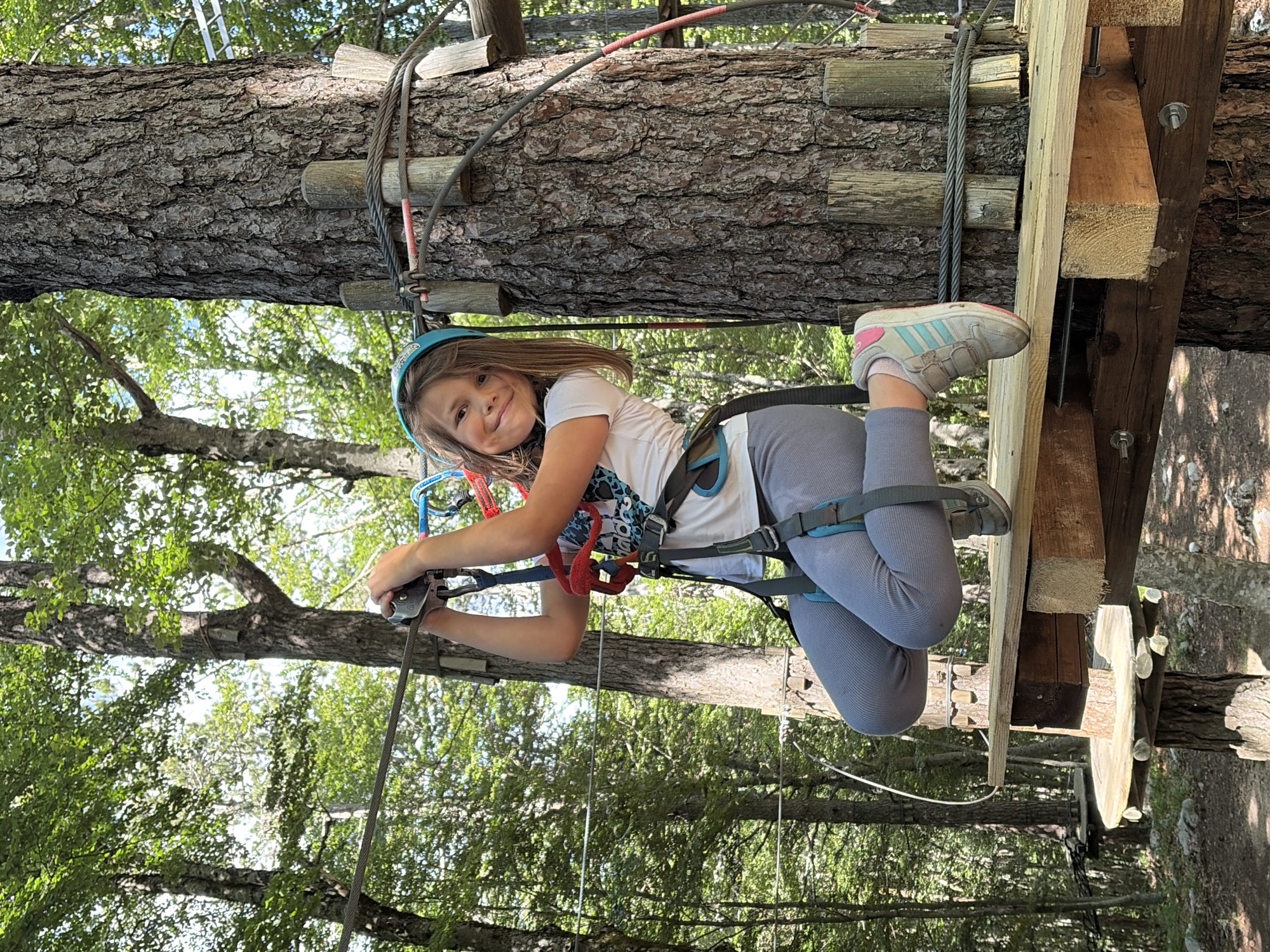 A young girl is depicted mid-air, suspended by a harness and ropes, participating in an adventure park activity. The image is oriented sideways, with the girl looking directly at the camera with a cheerful expression. She is wearing a light blue helmet, a white short-sleeved t-shirt with a graphic design, grey leggings, and grey and pink sneakers. Her body is angled, and her hands are holding onto a metal mechanism attached to a thick metal cable.
The scene is set outdoors, likely in a forest or treed area, suggested by the numerous tree trunks and green foliage visible in the background. A large tree trunk, which appears to be part of the adventure course structure, runs horizontally across the upper part of the image, with wooden planks and metal cables attached to it. Another wooden structure, possibly a platform, is visible to the right of the girl, also integrated with the tree. The sunlight filtering through the leaves suggests it is daytime with clear weather.
The girl's harness, secured around her waist and thighs, and the associated safety equipment, indicate she is engaged in a climbing or ziplining activity. Her smile conveys enjoyment and excitement. One of her sneakers shows the Adidas brand logo with three stripes. The activity appears to be taking place high above the ground, given the perspective looking down through the trees.