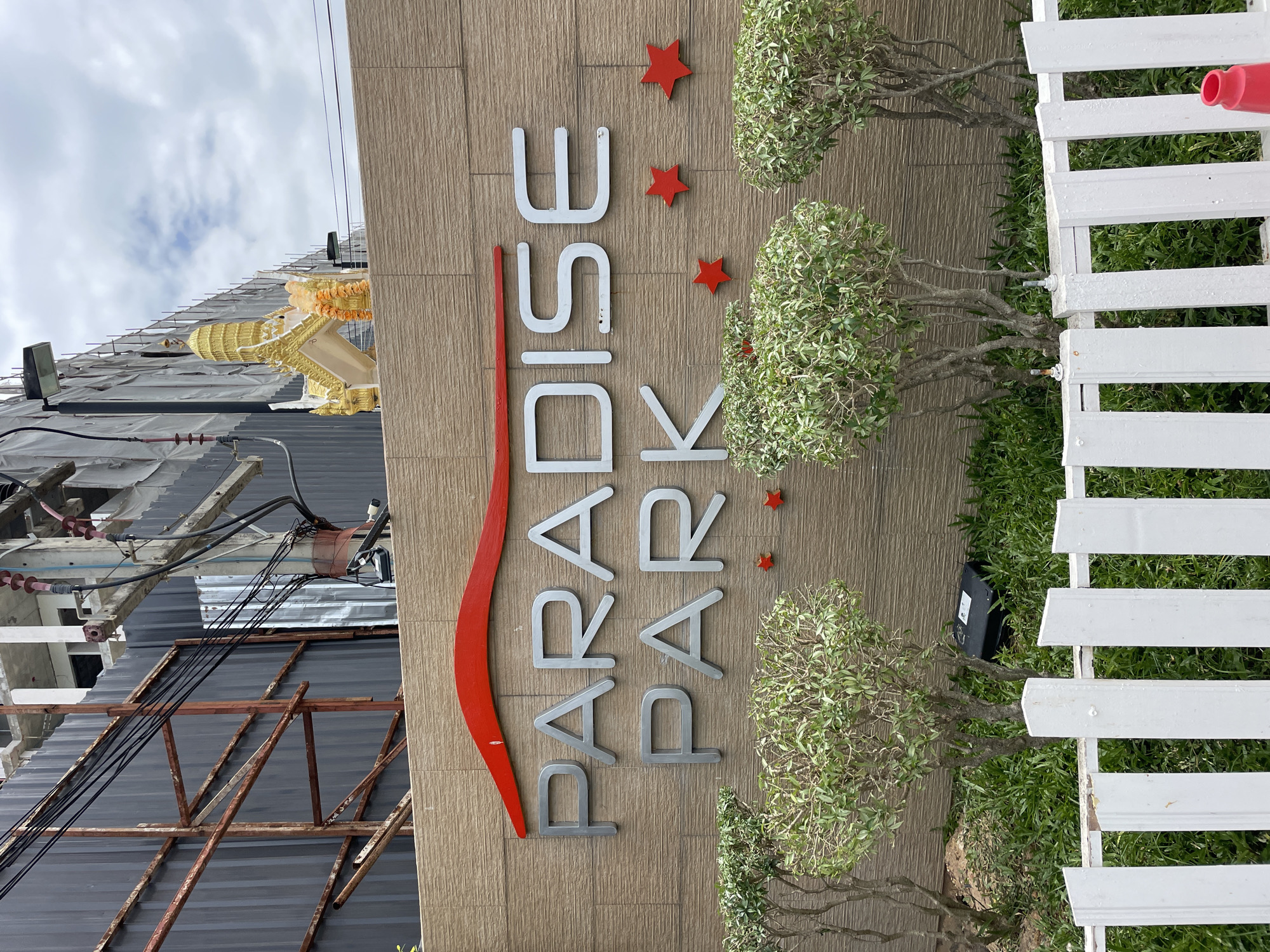 The image shows a sign for "PARADISE PARK" in Pattaya City, Thailand. The sign is made of silver metal letters mounted on a textured brown wall. Above the letters, there is a red wavy line. In the background, there is a building under construction with scaffolding and grey sheeting. A golden decorative structure, possibly a shrine or roof element, is visible at the top of the wall to the right of the sign. In front of the sign, there are several small, manicured green bushes. A white picket fence runs across the bottom of the image, with some of its posts appearing worn or damaged. A red traffic cone is visible to the right of the fence. The overall environment suggests an outdoor setting in a city, likely a commercial or residential area given the name of the establishment.