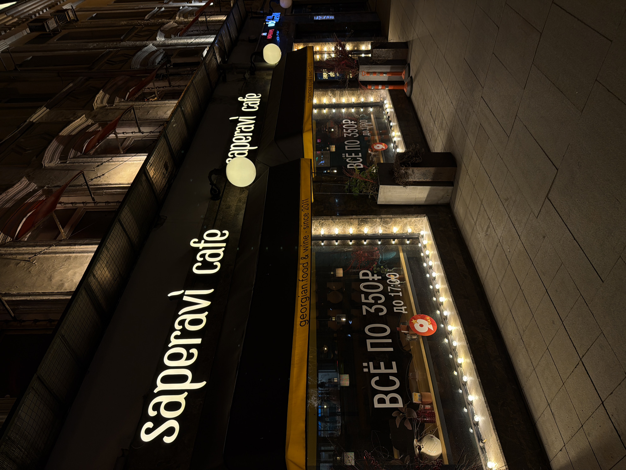 The image shows the exterior of "Saperavi Cafe" in Moscow, Russia at night. The cafe's lit signage is prominently displayed, advertising Georgian food and wine. The windows are outlined with strings of lights, and signs in Russian, advertising that everything is 350 rubles until 5:00 PM. The sidewalk is paved with large stone tiles. There are orange pylons/traffic cones in front of the café. The architecture above the cafe is visible, showing a multi-story building.