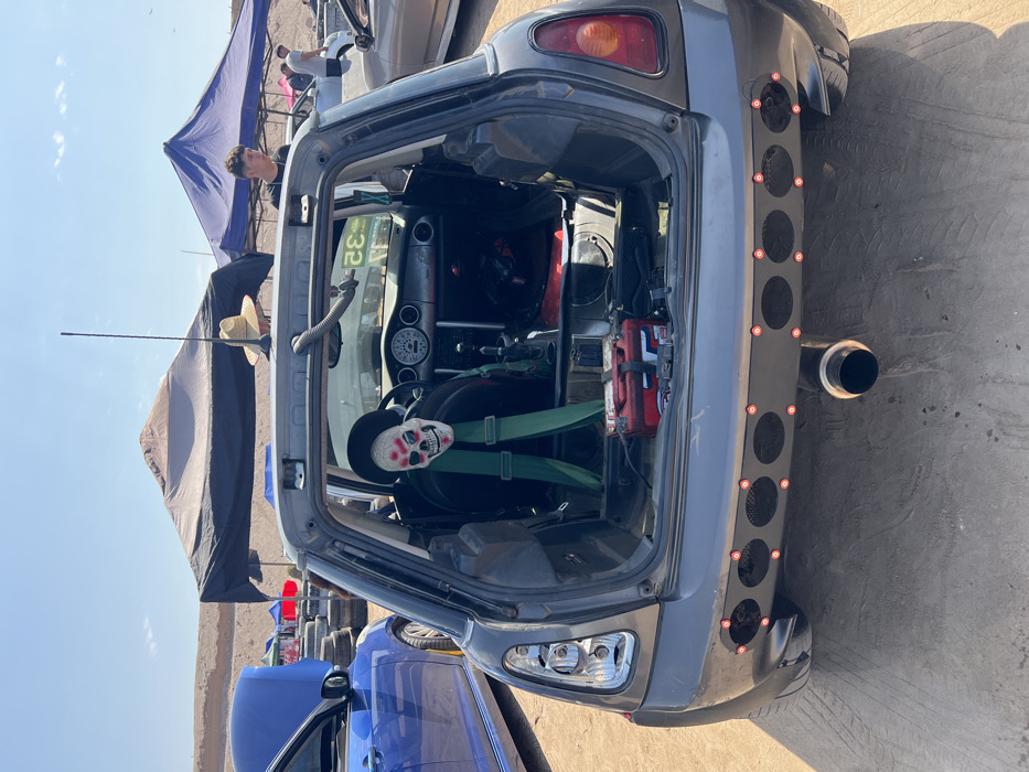 A gray Mini Cooper is angled to the left. The back is open, revealing a gutted interior with a skeleton figure strapped into the driver's seat with green racing straps. A red fire extinguisher is mounted to the right. The rear bumper has been replaced with a flat black bar with a row of round holes and a large exhaust pipe. Two tents are visible in the background, along with several other cars.