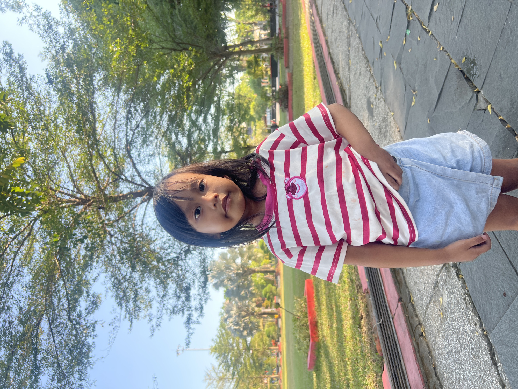A young girl with dark hair stands in a park, looking towards the camera. She is wearing a pink and white striped t-shirt with a cartoon character on it, and a light blue denim skirt. The girl is standing on a paved walkway. Behind her, a grassy area is visible with red-bordered planters and scattered trees. The sky is clear and blue, suggesting it is daytime. The overall scene depicts a relaxed outdoor setting, likely in a city park. The image appears to be taken in Purwodadi, Indonesia, given the distinctive palm trees visible in the background.