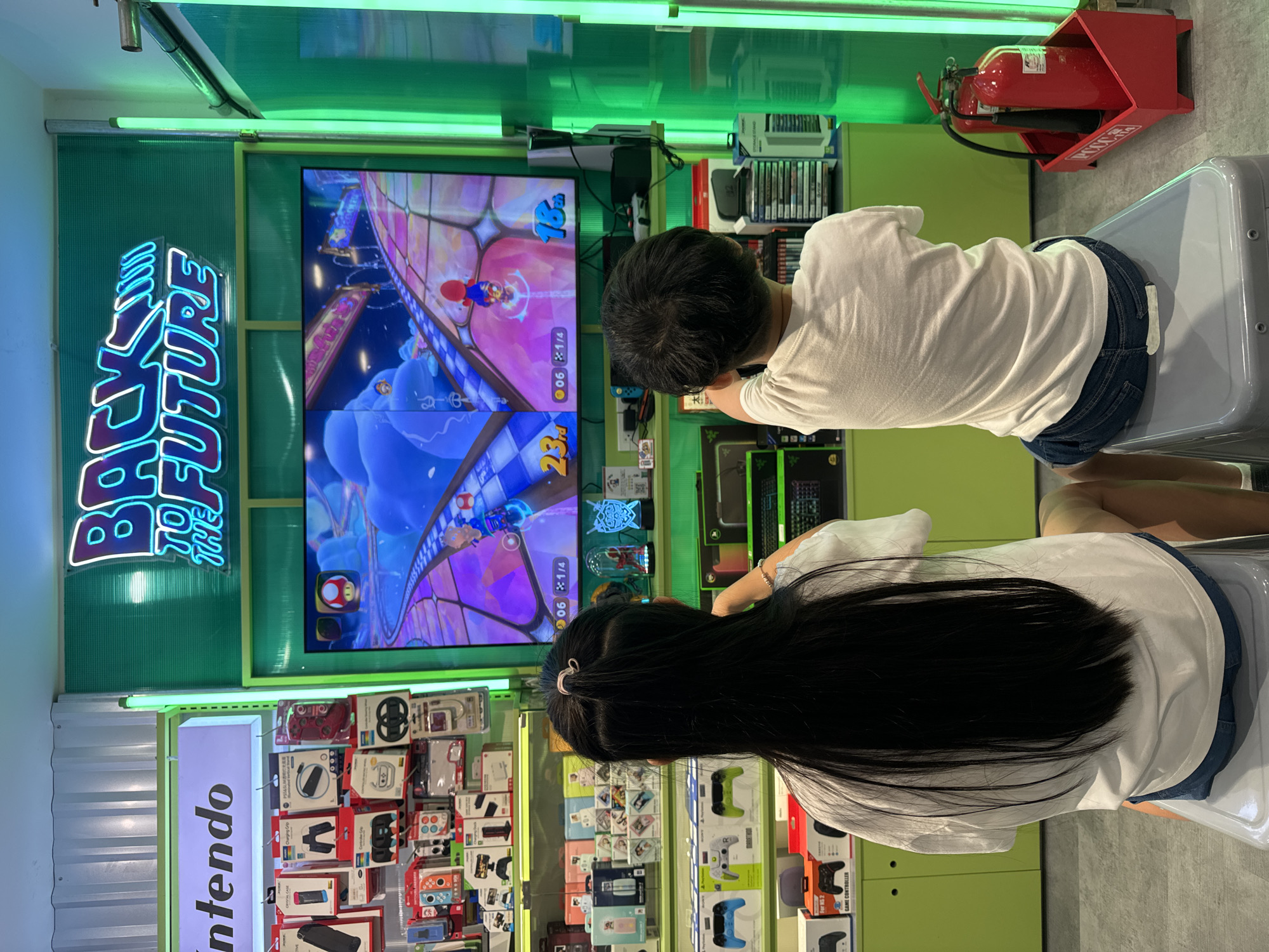 A child with dark hair and a white shirt is sitting in front of a screen playing a video game, likely a racing game such as "Mario Kart" due to the graphics shown on the screen, beside a person with long dark hair that is clipped back. The store, likely in Thủ Đức, Vietnam, features a "Back to the Future" neon sign, and a display of Nintendo products, games and accessories. A red fire extinguisher sits in a red container to the right of the child.