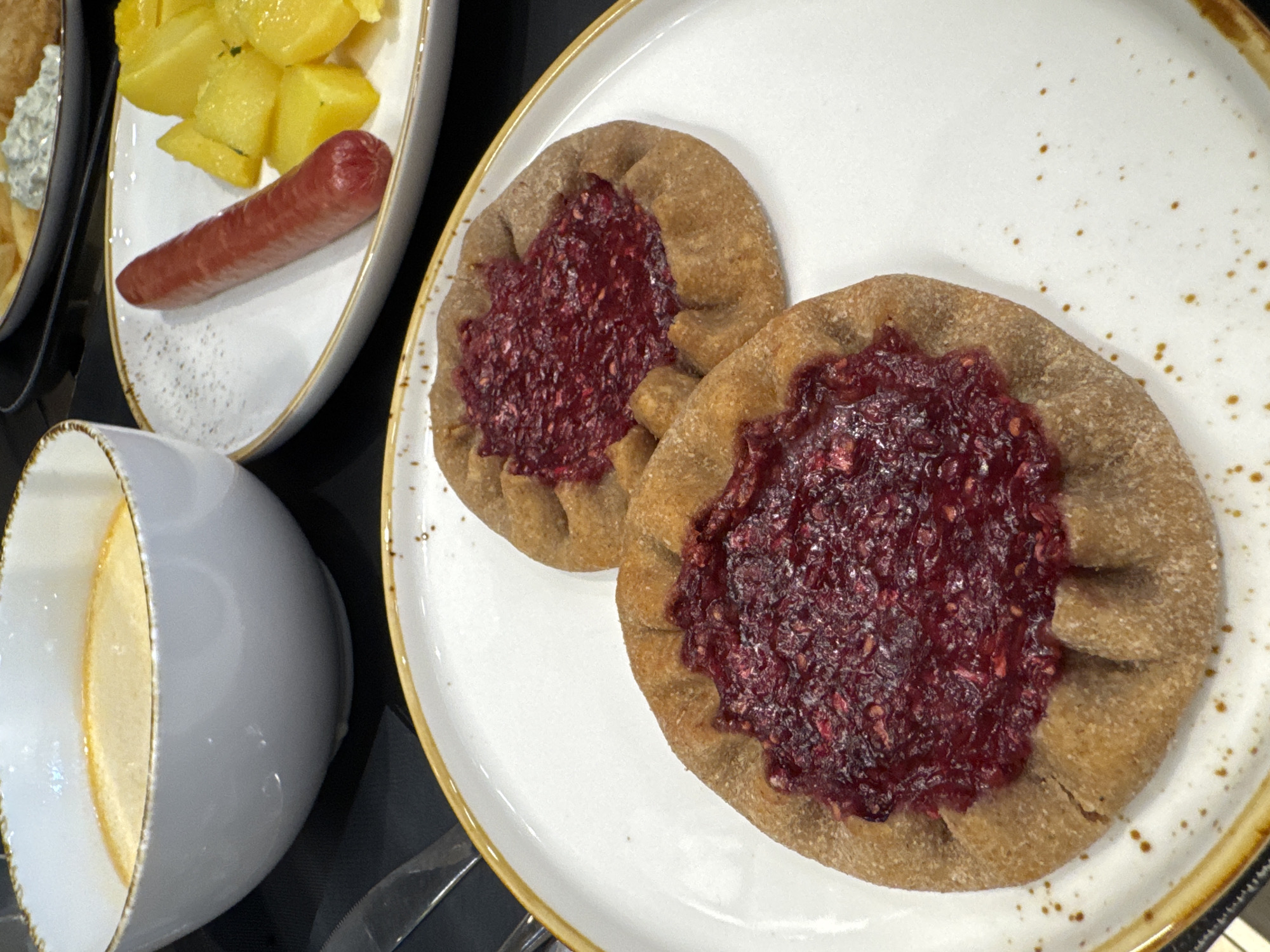 The image displays a close-up view of a meal laid out on a dark surface. In the foreground, two small, rustic pies with a golden-brown crust and a vibrant red berry filling are positioned on a white plate with a gold rim. The pies have a crimped edge, and the filling appears to be crushed berries, possibly raspberries.
To the upper left, a white bowl with a gold rim contains a light-colored soup or stew, with a slightly glossy surface.
In the upper right quadrant of the image, on another white plate with a similar gold rim, a single red sausage is placed next to several cubed, yellow potatoes. A small portion of what appears to be a creamy dip or sauce is visible behind the potatoes.
The overall setting suggests a meal served in a restaurant or home, with the focus on the food items. The Yakkima, Russia location is not visually discernible from the provided image.