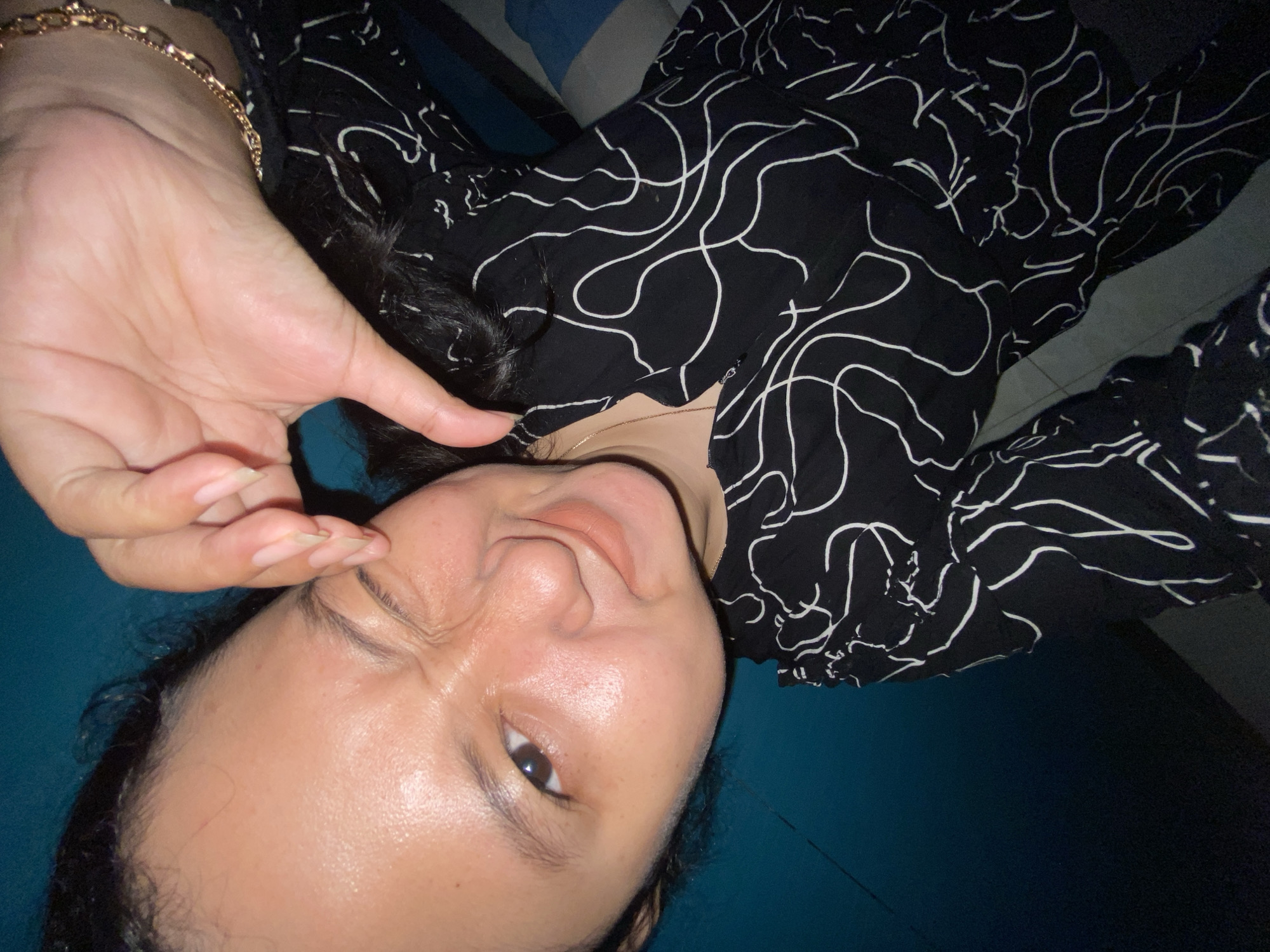 A selfie features a young woman with dark hair and brown eyes, winking and forming a heart shape with her fingers. She is wearing a black dress with a white abstract pattern and a gold bracelet on her wrist. The background is a gradient of dark blue. The image is cropped to focus on the woman's face and upper body. No specific location in Gelam, Indonesia is identifiable.