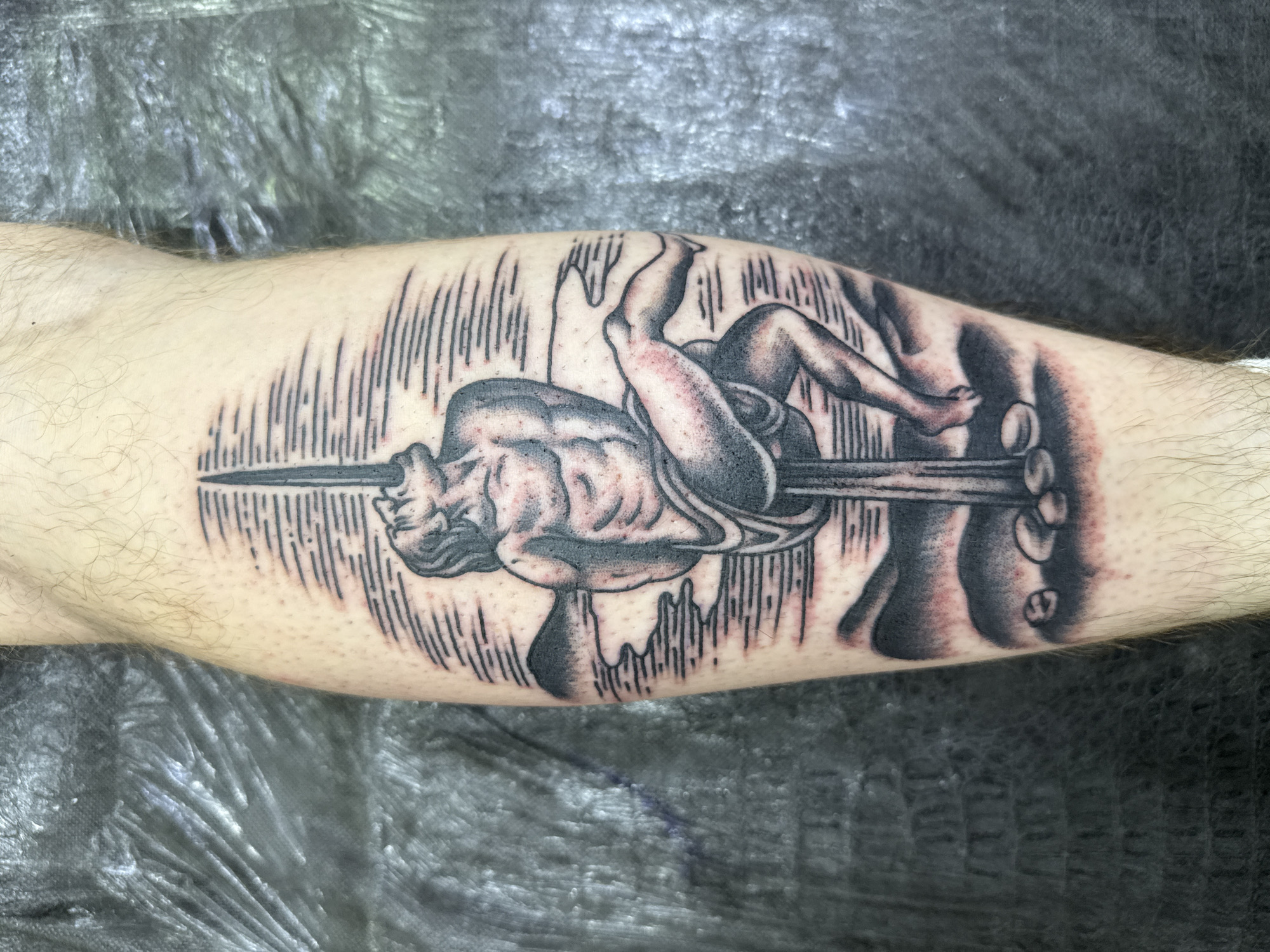 A tattoo on a person's calf depicts a man bound to a pole with a spear entering his mouth. The scene is rendered in a black and grey, woodcut style, with horizontal lines representing the background. The man's body is muscular, and his head is thrown back in apparent agony or ecstasy. He is seated with his legs bent. At the base of the pole are several stones. The background shows hills and water. The tattoo is located on the leg of an individual in Moscow, Russia.