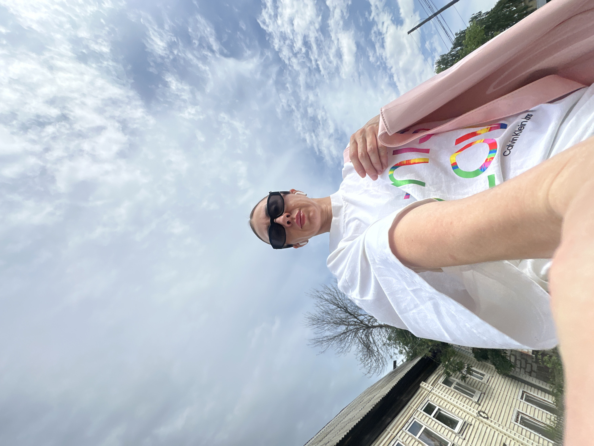 A selfie, angled upward, shows a person wearing dark sunglasses, a white T-shirt with rainbow colors and "Calvin Klein" printed on it, and a light pink jacket. The person is facing the camera and has one hand near their chest.
The background consists of a cloudy sky and parts of buildings and trees. A gray roof and a two-story house with multiple windows are visible. The location appears to be a residential area, potentially in Votkinsk, Russia.
