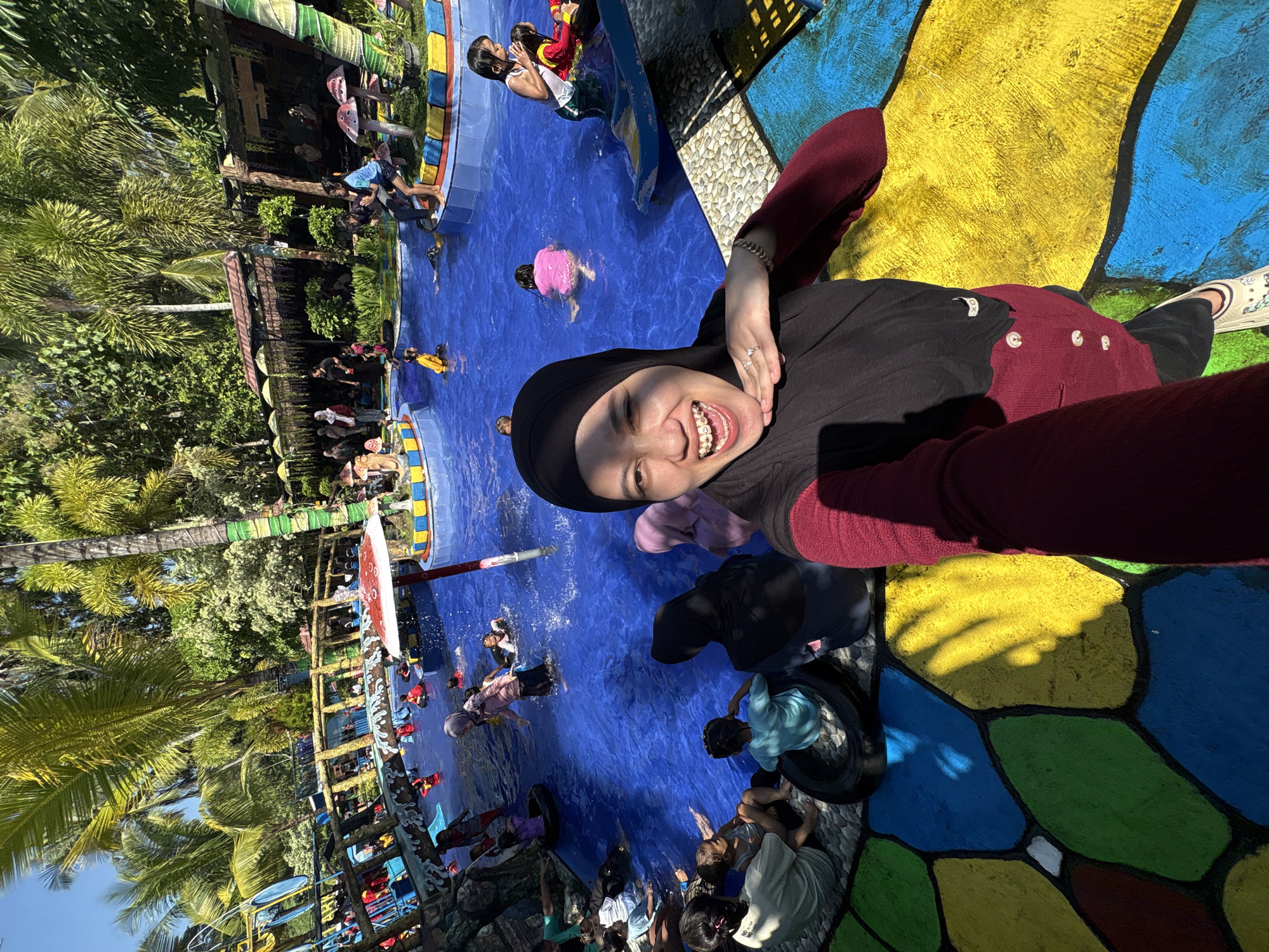 In Kebonsari, Indonesia, a woman wearing a black hijab and a maroon top is taking a selfie at a water park. The woman is smiling and has braces on her teeth. She is standing on a colorful, paved area with a geometric pattern of blue, yellow, green, and red shapes. Behind her, a vibrant blue pool is filled with people, mostly children, enjoying the water. Some people are swimming, while others are sitting on the edge of the pool. In the background, there are palm trees and lush greenery, typical of a tropical environment. There are also some playful mushroom-shaped decorations and a small bridge over the pool. The overall atmosphere appears to be joyful and recreational, under a bright, sunny sky.