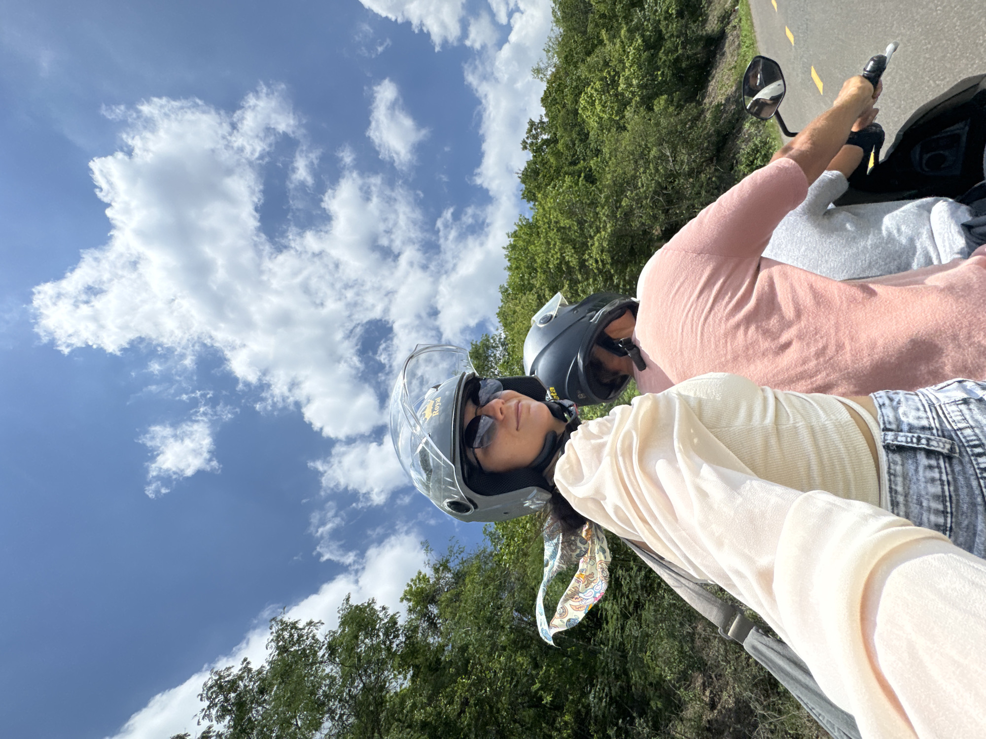 Two individuals are riding on a motorcycle on a paved road surrounded by lush green trees under a partly cloudy blue sky. The person in the foreground, wearing a light-colored, sheer blouse, denim shorts, and a grey helmet with a clear visor, is looking to the side. A colorful scarf is tied around their neck and flutters in the wind. They are wearing sunglasses and appear to be taking a selfie. The second person, in the background, is wearing a pink t-shirt and a black helmet and is operating the motorcycle. The road has yellow markings. The scene suggests a journey through a rural or natural area, possibly in the vicinity of Ham Thuan Bac, Vietnam, given the tropical foliage.