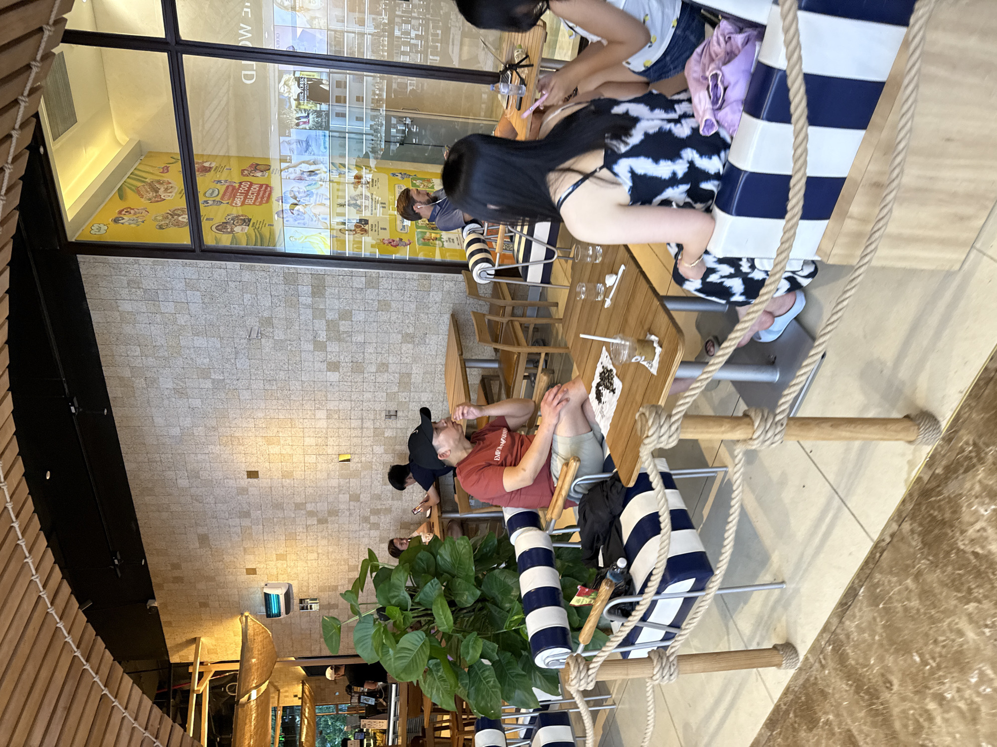 The image depicts the interior of a cafe in Nha Trang Ward, Vietnam. The scene is set with light-colored tiled walls and a wooden ceiling structure. Several patrons are seated at tables, engaged in various activities. In the foreground, a man in a red t-shirt and khaki shorts is seated at a table, looking down and eating. Adjacent to him, a woman with long black hair wearing a black and white patterned dress is seated, facing away from the camera. A child is seated next to her. Several tables and chairs are visible throughout the cafe, some occupied and some empty. A large potted plant is located near the center of the image. The cafe appears to be modern and casual, with rope barriers defining seating areas. The overall atmosphere is one of a relaxed dining environment.