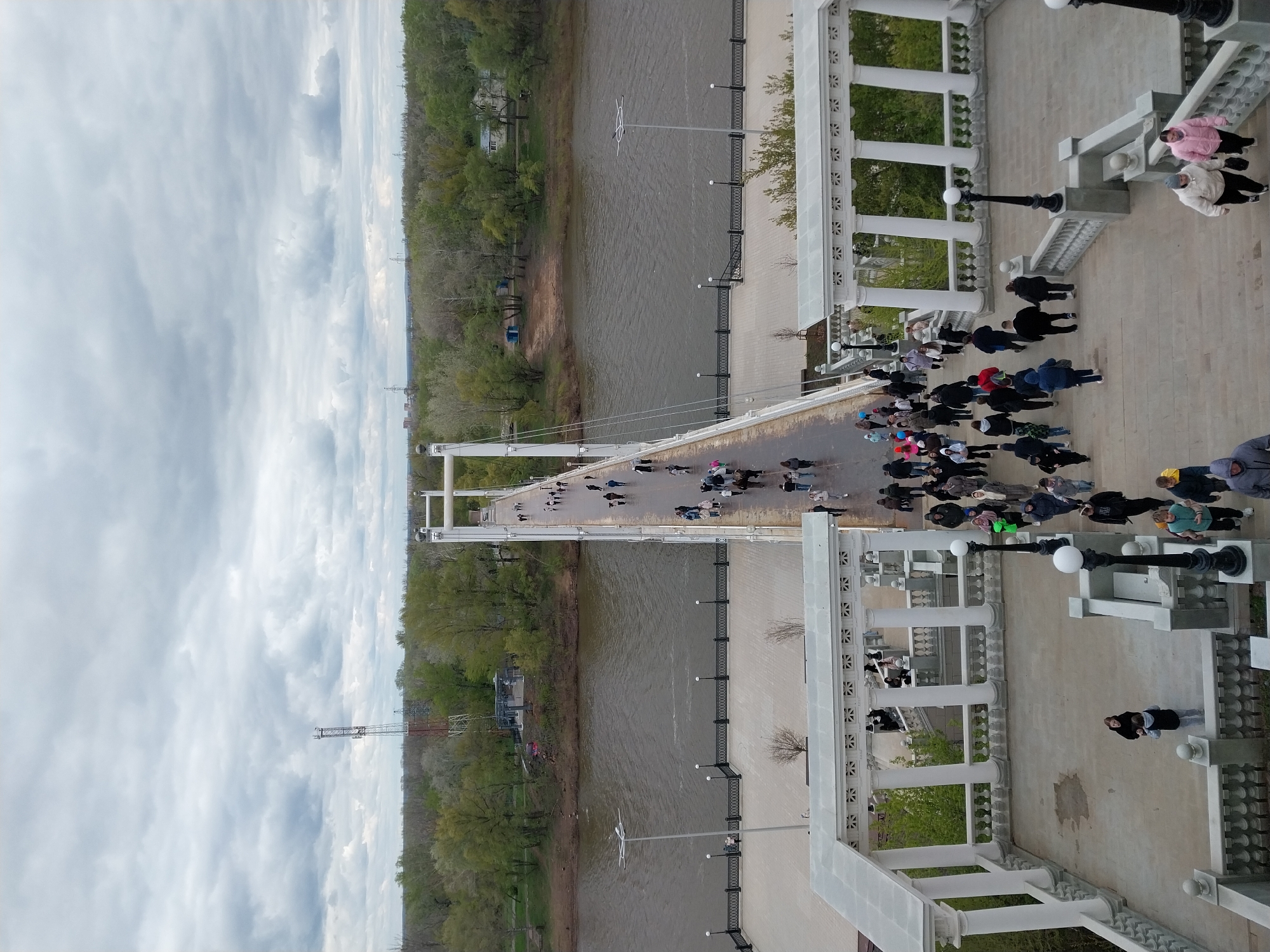 The image depicts a high-angle view of the pedestrian bridge over the Ural River in Orenburg, Russia. The bridge is a suspension structure with white towers. A group of people is gathered on the promenade near a white colonnade structure. Individuals are walking on the bridge. The Ural River is visible between the bridge and the riverbank. The sky is overcast with clouds. Trees line both sides of the river. A tall communication tower is visible along the riverbank. Streetlights are present along the promenade.