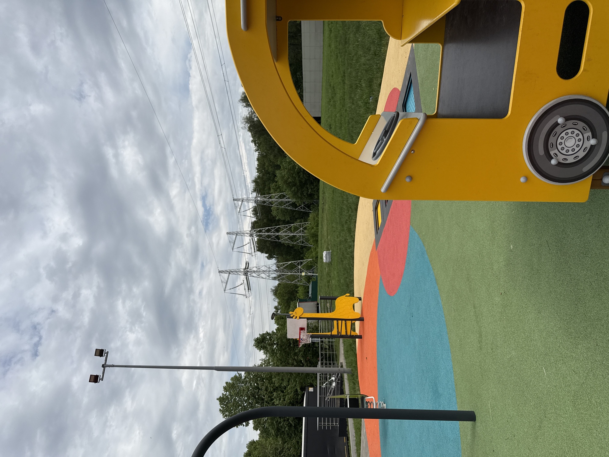 The image depicts a playground with various structures, including a yellow play vehicle and a basketball hoop with a giraffe-themed backboard. The ground is covered in brightly colored circular sections of what appears to be artificial turf.
In the background are power lines and pylons, with trees visible beneath them. A light pole is also present. The sky is overcast with gray clouds.
The location is possibly in Moscow, Russia.
