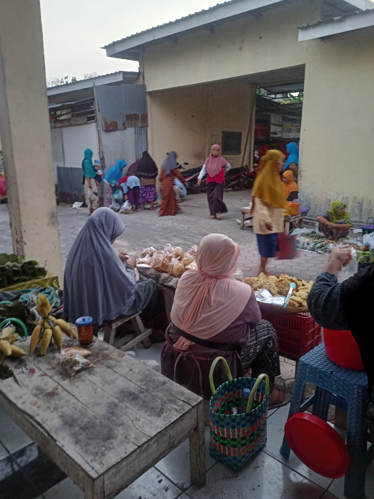 The image depicts an outdoor market scene in Nganti, Indonesia, likely during the daytime due to the ambient light. The foreground features a weathered wooden table laden with a bunch of yellow bananas, a jar with a blue lid, and small packets of what appear to be snacks. To the right, a woman wearing a pink headscarf and a dark skirt is seated on a wooden stool, her back to the camera, facing a display of fried food items arranged in a red plastic crate. Adjacent to her, a blue plastic stool is visible with a red lid resting beside it. Further back, several other individuals, predominantly women in headscarves and traditional clothing, are engaged in market activities. Some are interacting with each other, while others appear to be tending to their stalls. Stalls with various goods, including bags of food items and what could be produce, are set up. In the background, a building with a light-colored facade and a corrugated metal roof is visible, suggesting a permanent or semi-permanent market structure. The ground is paved with interlocking concrete blocks. The overall atmosphere suggests a bustling local market environment.