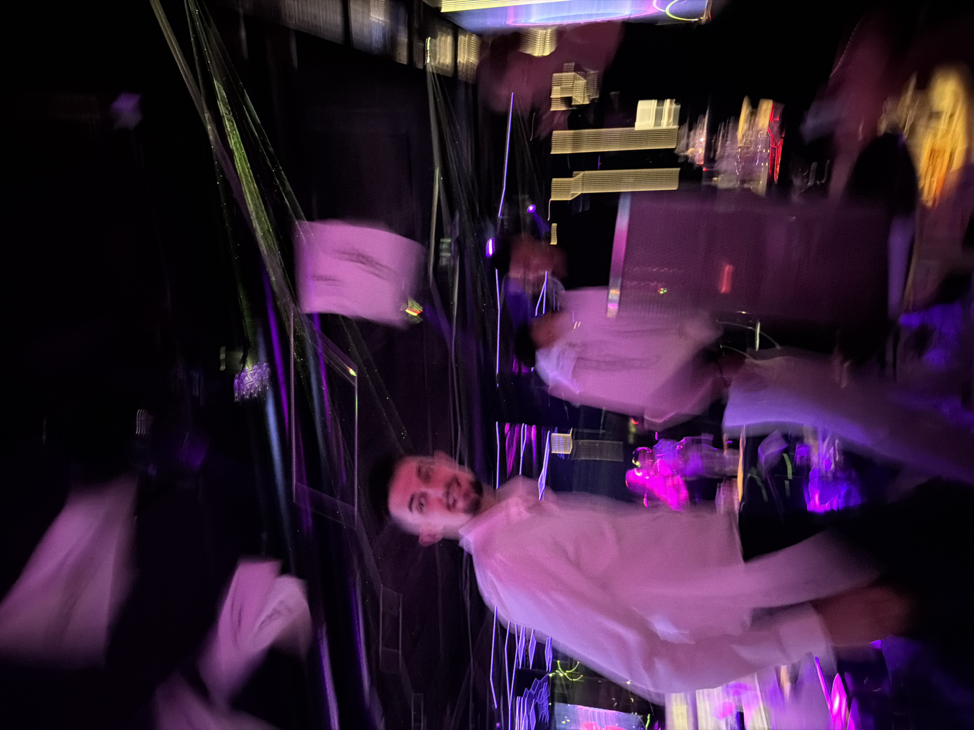 The image appears to be taken inside a dimly lit venue, possibly a nightclub or bar in the city of Sofia, Bulgaria, based on architectural characteristics and cultural context. The photograph is blurred and exhibits motion, indicating low lighting conditions or camera movement. There are multiple individuals present, but their features are indistinct due to the blur. One individual, a male, is wearing a white shirt and seems to be the focal point, although he is not clearly in focus. The background contains vertical lighting elements in purple and green, adding to the ambiance. It looks like a social gathering or a celebratory event, with people possibly interacting with each other, though the details are vague due to the image quality. Objects include barware and other equipment, but these are not clear either.