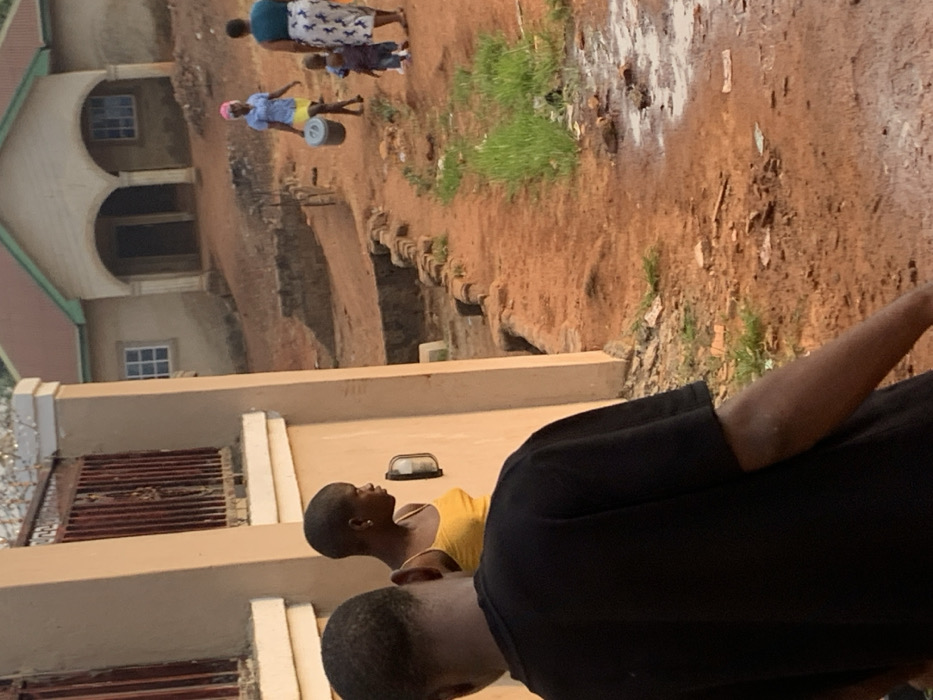 This outdoor scene, set in Otuocha, Nigeria, captures a glimpse of daily life. The ground is primarily reddish-brown earth, uneven in places, with sparse patches of green grass and some lighter, possibly sandy or disturbed, areas.
In the background, a two-story building with light-colored walls and a green roof is visible. It features distinct arched entrances or windows on the ground floor and rectangular windows with white frames on both levels. To the left in the mid-ground, another building with concrete walls and a metal gate/fence is partially seen. A dirt path or slope with rough steps or retaining structures appears to lead down.
Several people are present. In the far background, a woman wearing a patterned skirt and a dark top is seen holding a small child by the hand. Further back on the left, another person, likely a woman, is walking while balancing a light grey or white bucket on her head. She is dressed in a light blue top, a yellow skirt, and a pink head covering. In the foreground on the right, a person with short dark hair, wearing a black t-shirt, is seen from the back and side. Slightly behind them, another individual with short hair, wearing a yellow top, is shown in profile, appearing to look upwards or to the side.
The scene depicts common activities like walking and carrying goods, suggesting routine daily life in a residential or semi-rural setting. The natural lighting indicates it is daytime, possibly late afternoon, with no harsh shadows. No text is visible in the image.