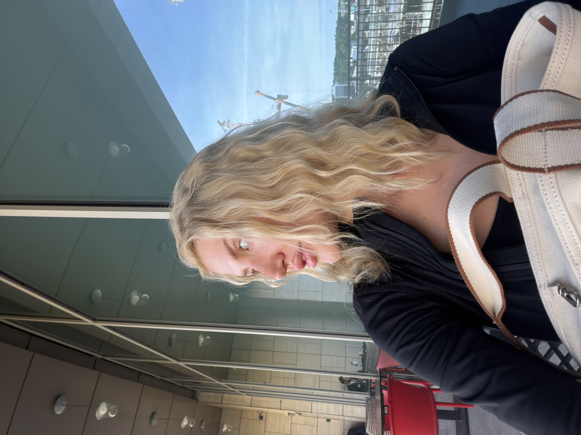 A medium shot, taken indoors, depicts a blonde-haired woman in profile. She is wearing a black jacket and a cream-colored canvas bag with brown trim is partially visible. The background shows a glass-paneled wall offering a view of a cityscape featuring cranes and buildings. Red chairs and tables are partially visible near the woman, along with other people reflected in the glass. The location is potentially in Belgrade, Serbia.