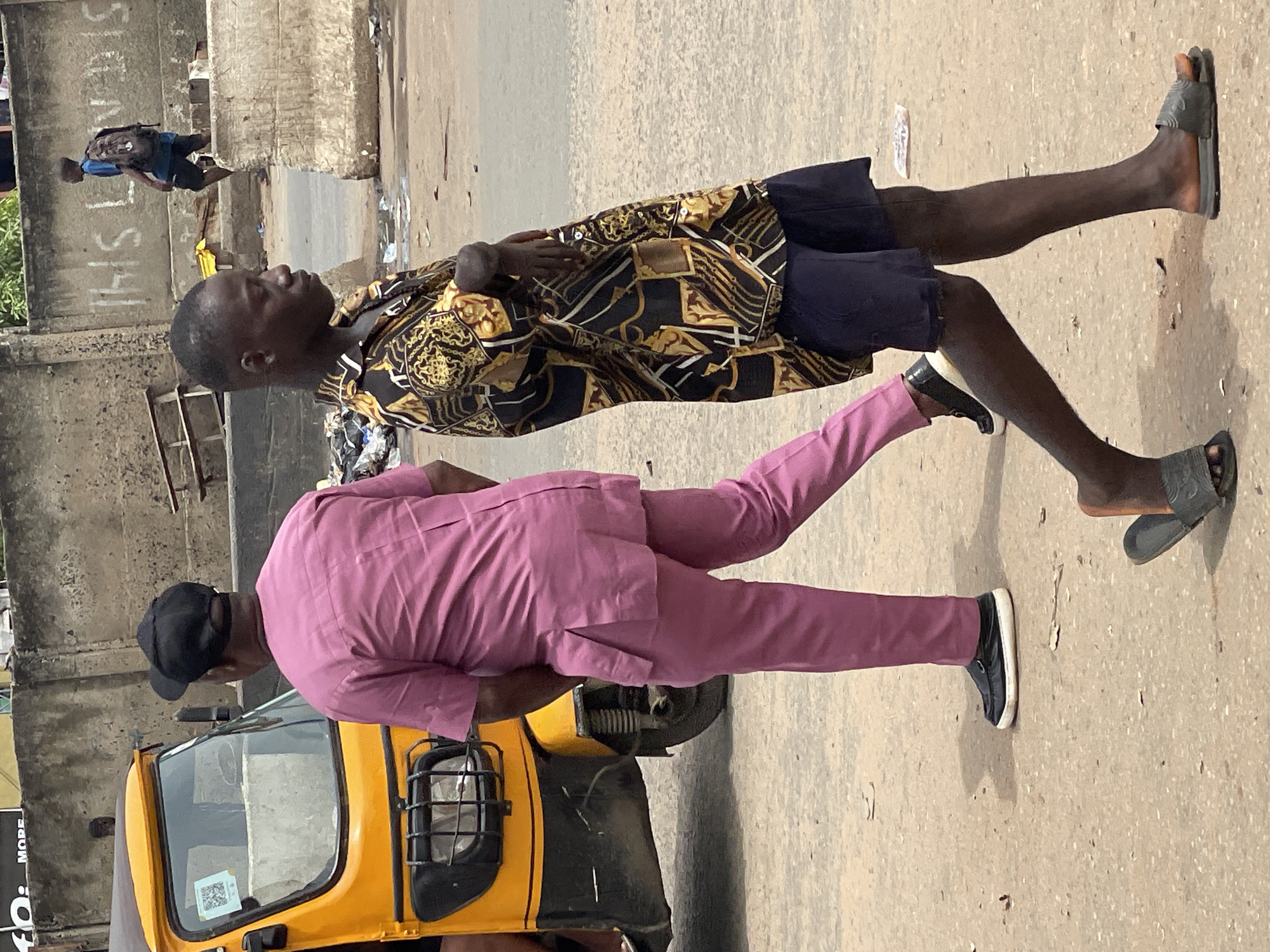 Two men are walking on a paved surface in Onitsha, Nigeria. The first man is wearing a black and gold patterned shirt, dark shorts, and grey sandals. The second man is wearing a pink suit and black shoes, and standing near a yellow tricycle taxi. In the background, there is a building with graffiti on the side. Another person with a backpack is walking in the distance. The environment appears to be an urban street.