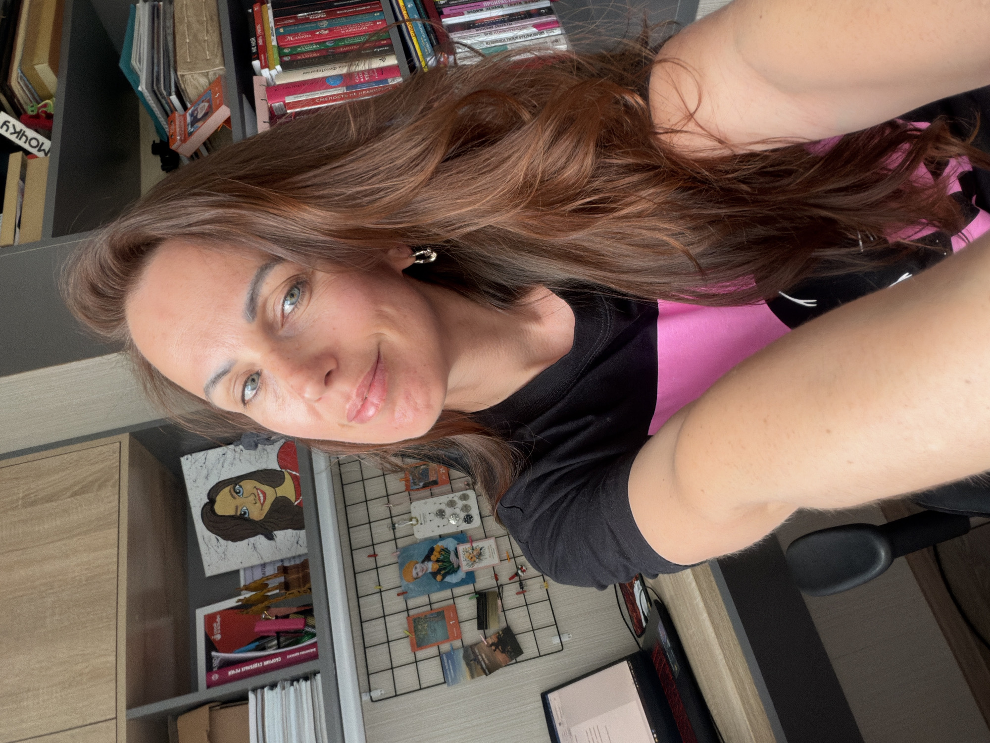A woman is captured in a selfie, looking directly at the camera with a slight smile. She has long, wavy brown hair, fair skin, and light-colored eyes. She is wearing a black t-shirt with a pink section and a silver-colored earring is visible in her right ear.
The setting appears to be a home office or study room in Birobidzhan, Russia, indicated by the text on the books and the provided location context. Behind her, a grey and wooden shelving unit is filled with numerous books, mostly in Cyrillic script. Above the woman's head, part of a shelf organizer reads "МОЙ" (MY). On the shelves, book titles or authors visible include "В. А. Каверин: ДВА КАПИТАНА" (V. A. Kaverin: TWO CAPTAINS), "СМЕЛОСТЬ И НАДЕЖДА" (COURAGE AND HOPE), "ВАДИМ ЗЕЛАНД" (VADIM ZELAND), "ЖЕНСКАЯ" (WOMEN'S), and "СБОРНИК СУВЕНЕВЫХ РЕЧЕЙ" (COLLECTION OF SOUVENIR SPEECHES).
To her right, a desk is visible with a computer or laptop screen showing text. Above the desk, there's a black wire grid-style pinboard adorned with several small photographs, postcards, and clips. A framed picture or painting of a cartoonish woman with brown hair and blue eyes hangs on the wall or shelf. The lighting suggests it is daytime.
