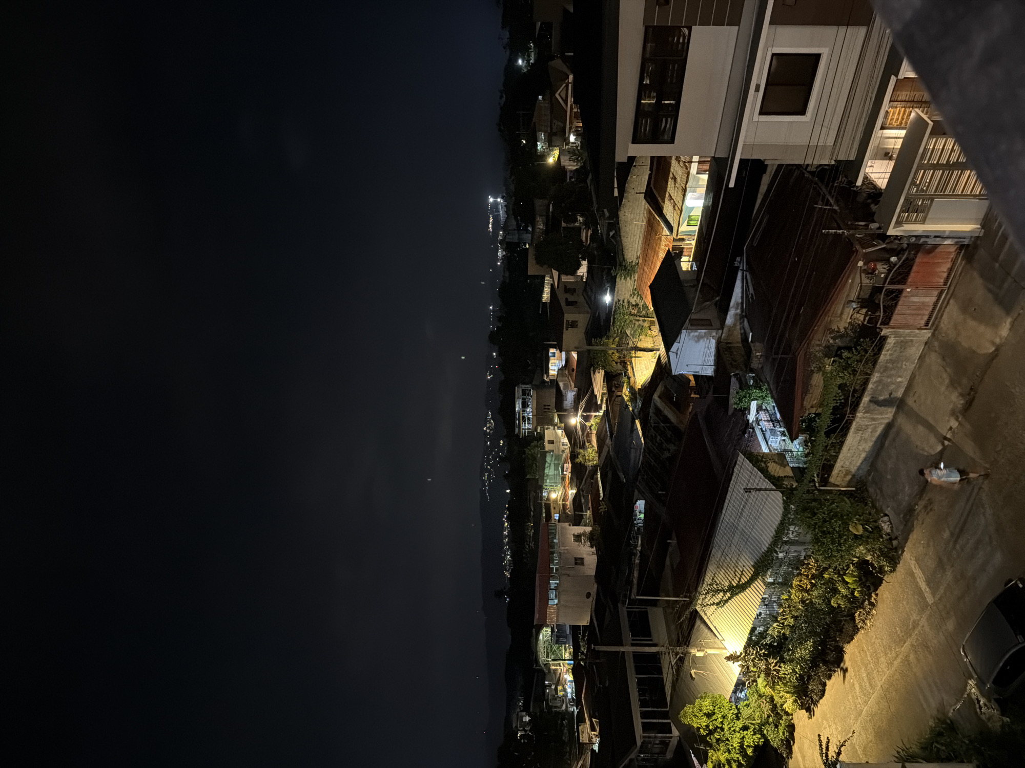 The image shows a nighttime view of a residential area in Davao City, Philippines. The right side of the image is dominated by densely packed houses with corrugated metal roofs and concrete structures. Streetlights illuminate the narrow streets, revealing lush vegetation and parked vehicles. On the left side, a dark skyline suggests a view towards the sea or open space. Dim city lights are visible on the horizon. In the lower right corner, a person is lying on the street.
