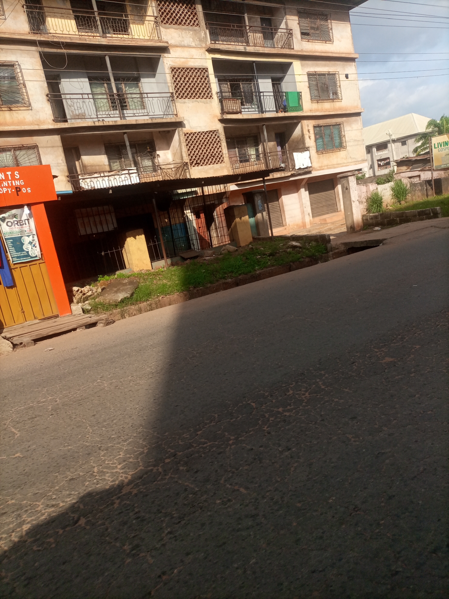 The image captures a street scene in Nkpor, Nigeria, with a focus on a multi-story building and the road in the foreground. The building appears to be somewhat aged, with a light-colored facade and balconies with metal railings. Some windows are covered with metal grates, and a few items like laundry are hanging from balconies.
In front of the building, there are several shops with closed shutters, suggesting it might be outside of business hours or a quiet period. An orange sign with white lettering is visible on the left, which appears to be advertising a business. One of the shops has an open area with some displays, including a poster with text. The ground in front of the shops is unpaved and overgrown with some vegetation, indicating a mix of urban and natural elements.
The road in the foreground is paved but shows signs of wear and tear, with a cracked surface. A shadow of an unseen object stretches across the road, hinting at the time of day, likely late afternoon or early morning with the sun low in the sky. The sky above is partly cloudy, suggesting fair weather.
In the background, another building with a lighter-colored roof is visible, along with some greenery and a sign that appears to be for "LIVING GOSPEL CHURCH". The overall impression is a typical street view in a Nigerian town, showcasing local architecture, commercial activity, and the urban landscape. No people are prominently visible in the scene, and the focus is on the static elements of the environment.
Visible text includes "NTS PAINTING COPY-POS" and "ORBIT DIGITAL PRINTS" on the orange sign and poster respectively. There is also text on the distant sign, part of which reads "LIVING GOSPEL CHURCH".