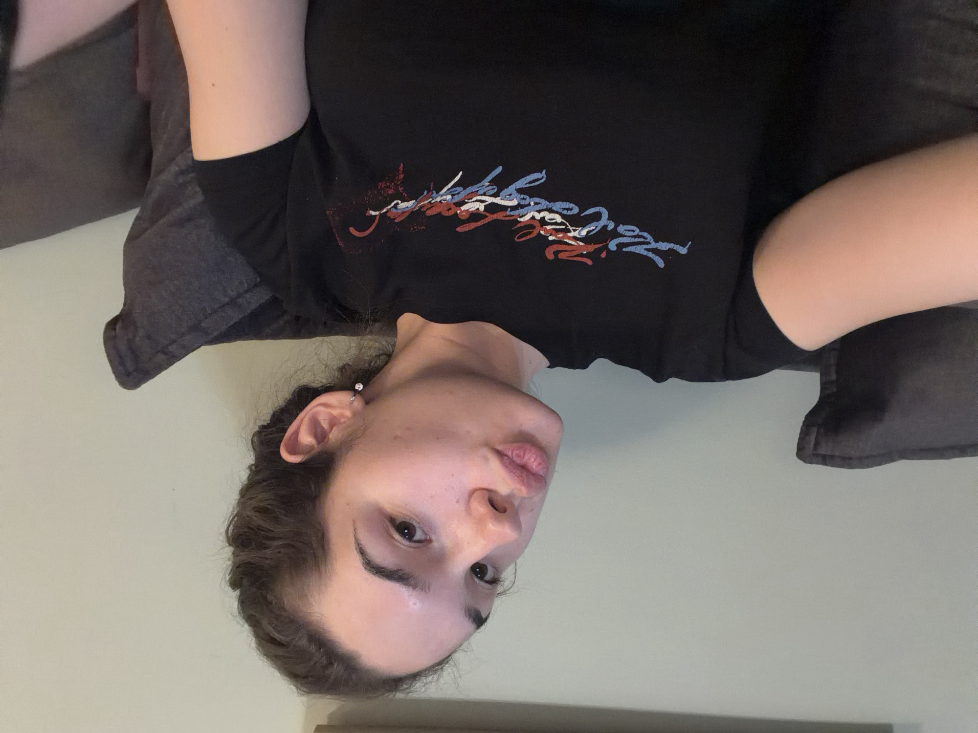 A woman is lying on a grey pillow with a black t-shirt on, featuring a red, white, and blue design. She is looking directly at the camera with a slight pout. There is a wall behind her, and a part of what looks like a wooden desk or shelf is visible at the bottom.