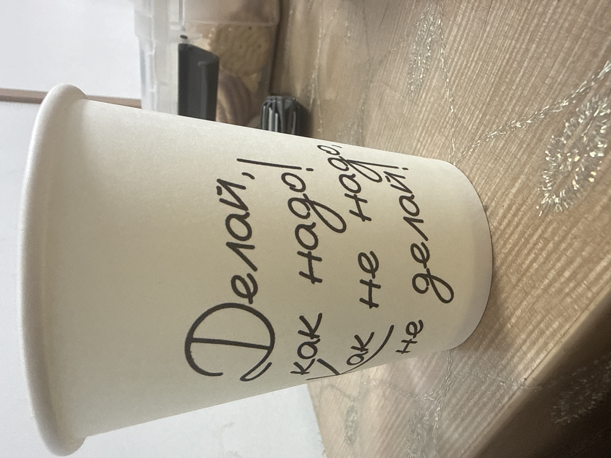 A close-up, eye-level shot shows a white paper coffee cup with handwritten black text. The text appears to be in Russian and reads "Делай, как надо! Как не надо, не делай!" which translates to "Do as you should! As you shouldn't, don't do!". The cup is positioned on a wooden surface that has a textured grain and some scattered shiny embellishments, possibly tinsel. The background is blurred, showing parts of what might be a shelf or counter with some containers, suggesting an indoor setting. The lighting is soft and diffused. There are no people visible in the image. The specific location in Beslan, Russia cannot be determined from the visual information provided.