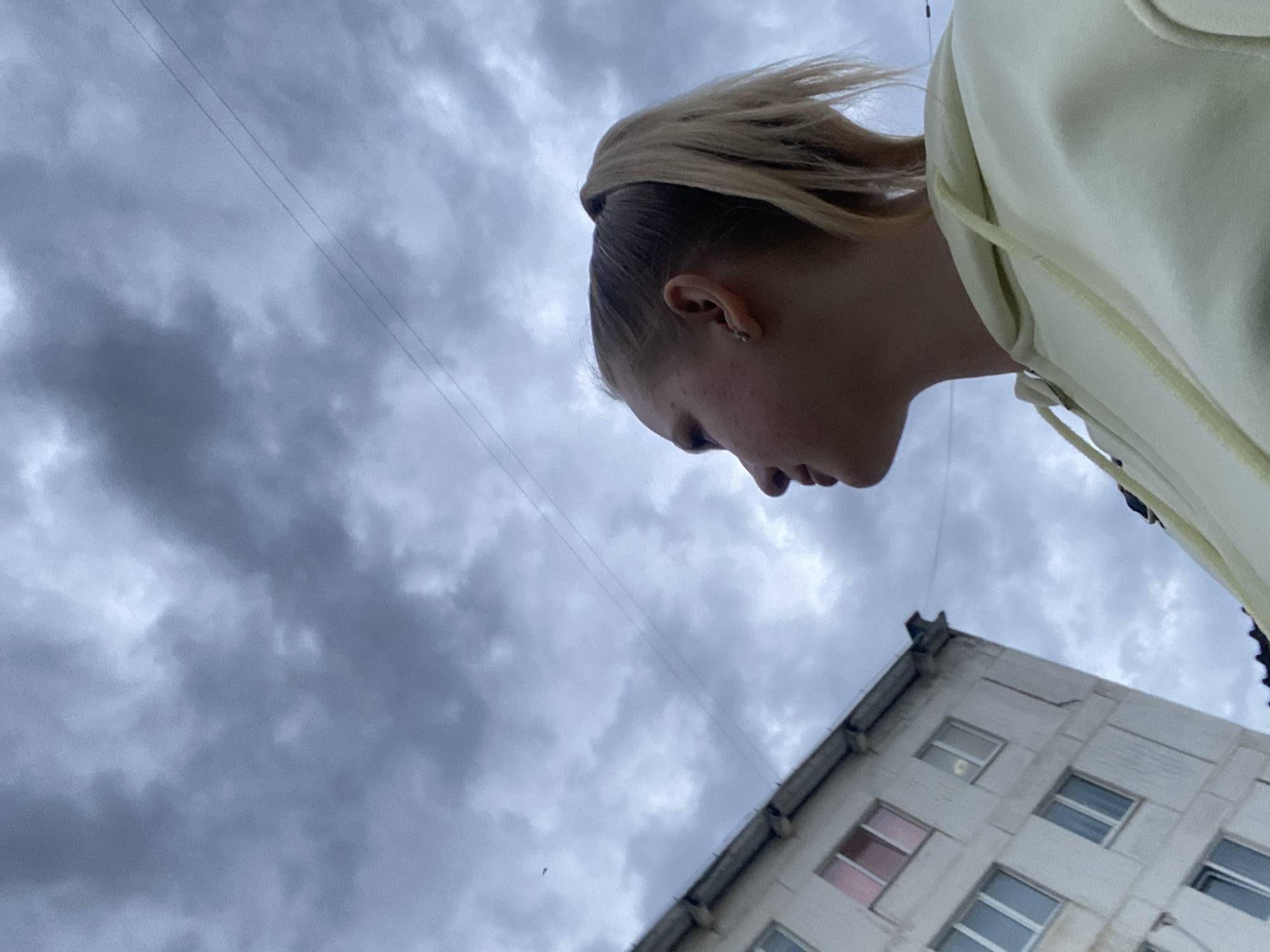 The image is a low-angle shot of a person with blonde hair in a high ponytail and wearing a light-colored hooded sweatshirt. The person's face is partially visible in profile. Behind the person is a cloudy, overcast sky. At the bottom right of the frame is a multi-story building with several windows, suggesting an urban environment. Thin power lines are visible against the sky. There are no explicit actions or interactions depicted. The image provides minimal information that allows for a definitive location. Chaikovsky, Russia is unable to be located due to limited information available.