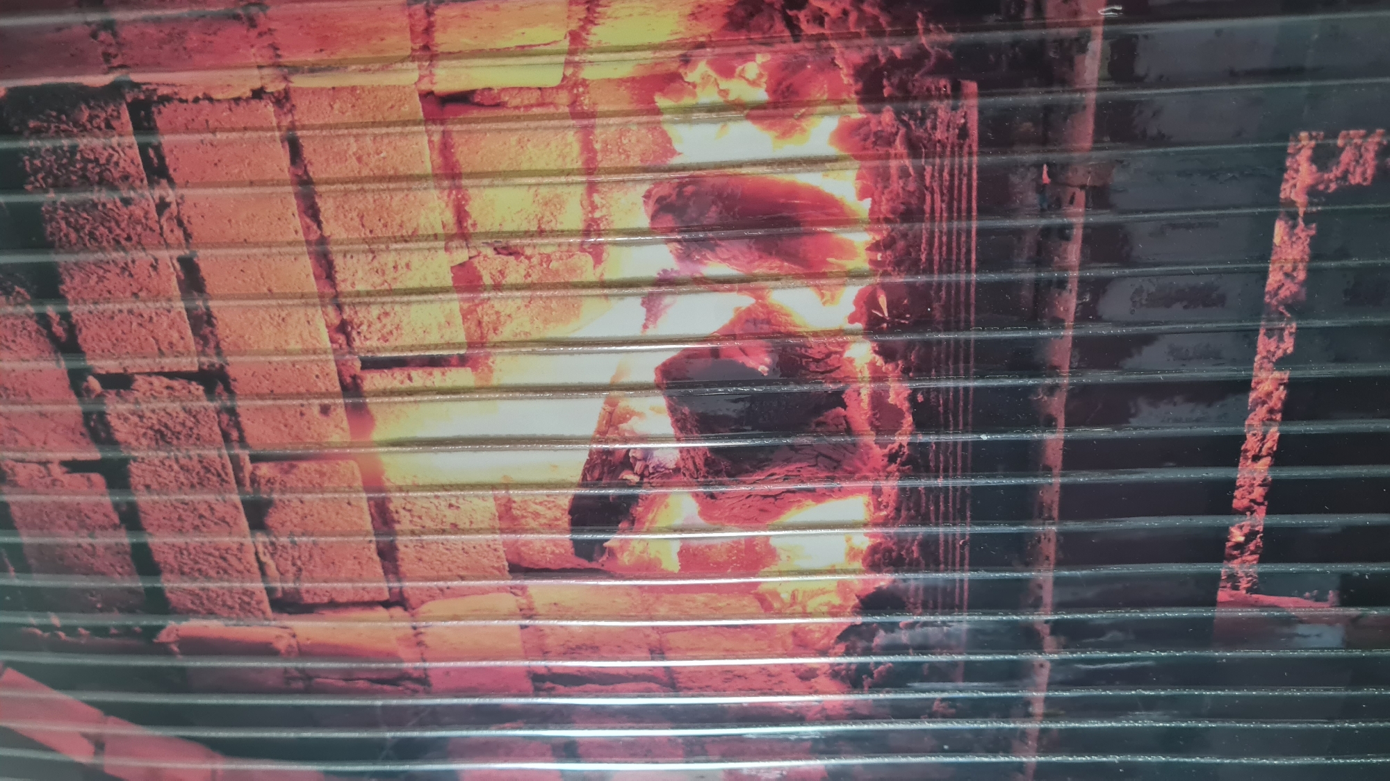 The image depicts a depiction of a fireplace behind horizontal bars. The fireplace is constructed of bricks and contains a fire. It is unclear what the image is printed on. There is no indication of the location in Nahariyya, Israel. No people, subjects, or interactions are visible.