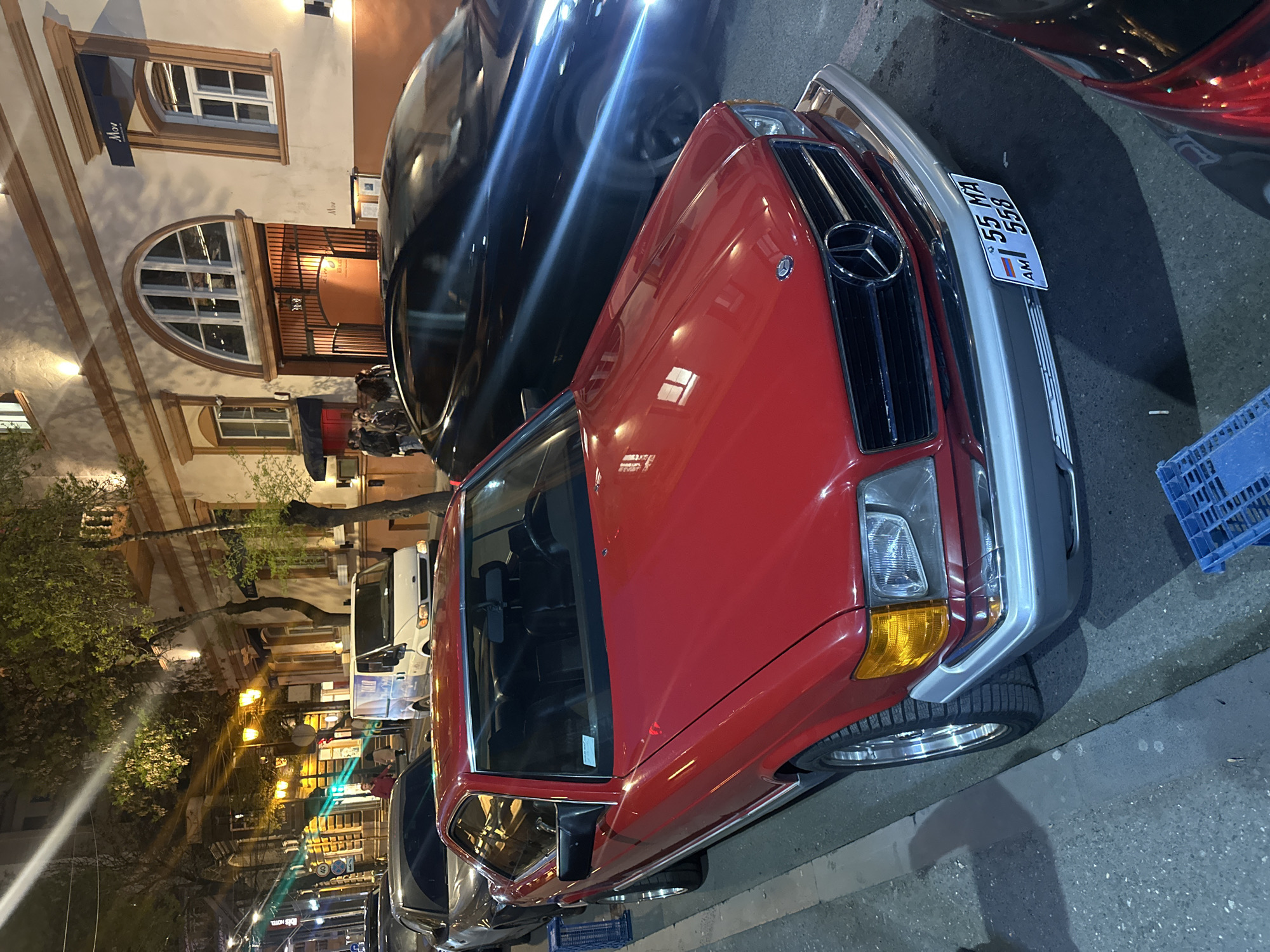 A red Mercedes-Benz, with license plate "AM 55 MA 558", is parked on a Yerevan, Armenia, street. A black car is parked next to it. The location is a street with shops and restaurants. Several pedestrians are standing near a business in the background. The streetlights are on.