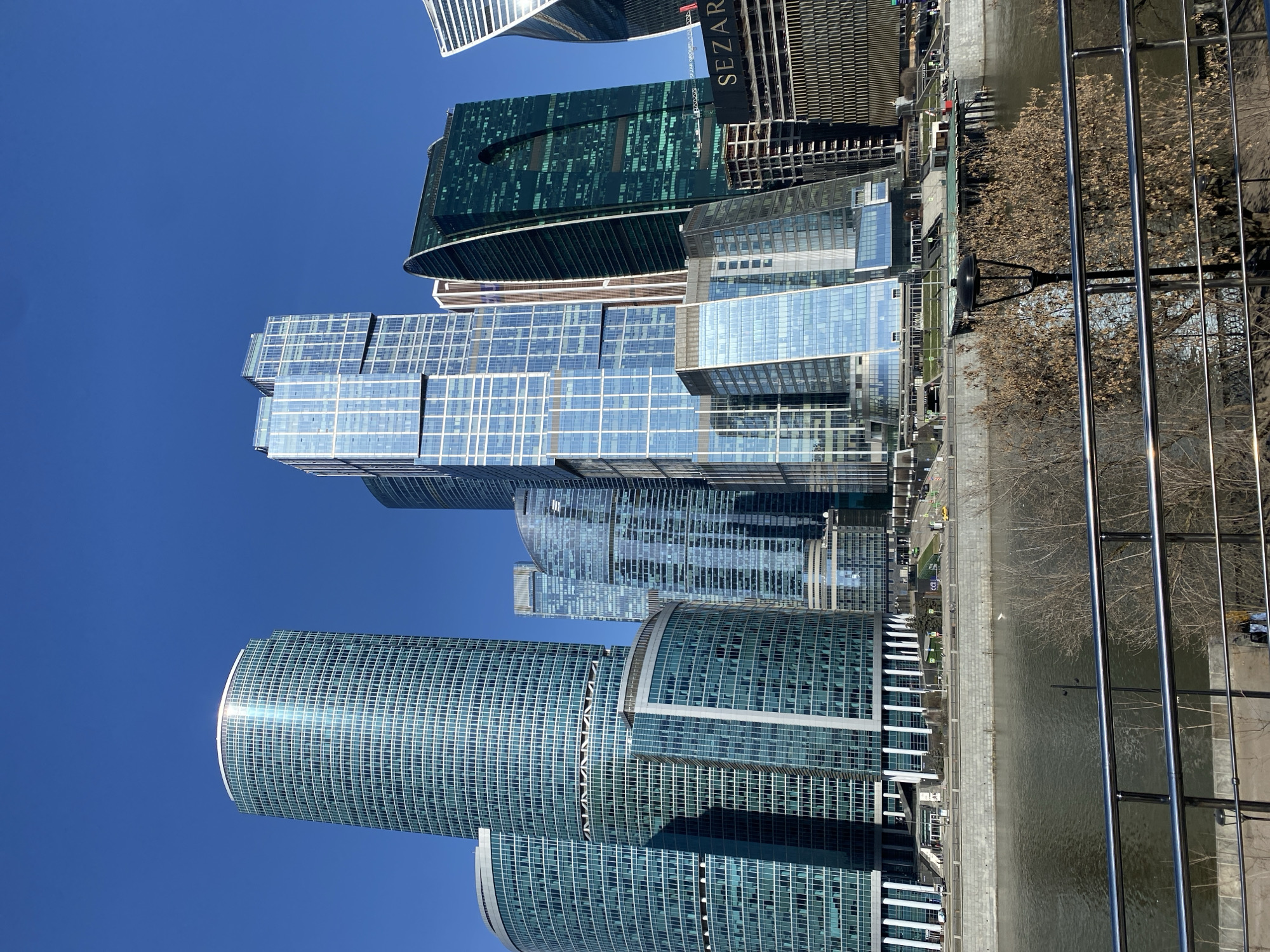 The image depicts the Moscow International Business Center (Moscow City) in Moscow, Russia, viewed from an elevated perspective. The scene showcases several modern skyscrapers constructed with glass and steel, reflecting the clear blue sky. A body of water, likely the Moskva River, runs along the right side of the frame, separated by a concrete embankment. Some trees with bare branches are visible along the embankment, indicating a time of year when leaves are absent. A metal railing is present in the foreground. There are no visible people.