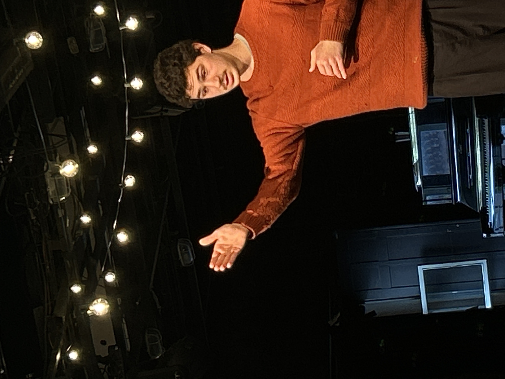 The image captures a young man in a dimly lit, possibly theatrical, setting. He has short, curly dark hair and a light complexion, wearing a rust-orange knitted sweater over a white shirt, and dark trousers. His expression is serious and direct, with a slight tilt of his head. He extends his right arm forward and slightly down, with his palm facing upwards, as if gesturing or offering something.
To the left of the man, a string of warm, glowing spherical lights hangs vertically, illuminating the dark background. These lights appear to be decorative, contributing to the ambiance of the scene. The background is predominantly dark, suggesting either night or a darkened interior space like a stage. On the far right, indistinct dark structures, possibly stage equipment or part of a building, are visible. In the lower right, a rectangular, light-colored frame with a dark interior is set against a dark surface, resembling a small window or a framed panel. The overall impression is one of a performance or a posed shot within a dramatic or artistic environment. There is no visible text in the image.