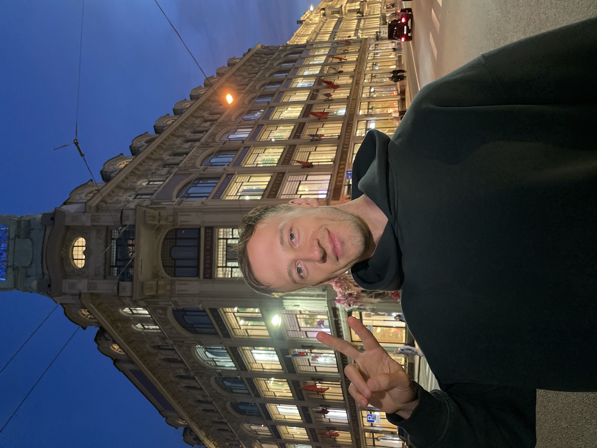 A medium shot photograph taken at dusk captures a man in the foreground, a building in the background, and a street scene. The man, fair-skinned with short brown hair and a light stubble, is wearing a dark green hooded sweatshirt. He is smiling and holding up his right hand with two fingers extended in a peace sign. Behind him, a grand, ornate, multi-story building with numerous lit windows dominates the frame. The building's architecture suggests a historical European style, characteristic of Saint Petersburg, Russia. The facade features intricate detailing, balconies, and arched windows. Flags are visible hanging from some of the windows. The street in front of the building is paved, with a car visible in the distance on the right side of the frame, its red taillights illuminated. The sky above is a deep blue, indicating twilight. The scene is illuminated by the lights of the building, streetlights, and the ambient light of dusk. The location is identifiable as the Singer House (Dom Knigi) on Nevsky Prospekt in Saint Petersburg, Russia, due to its distinctive architecture and signage.