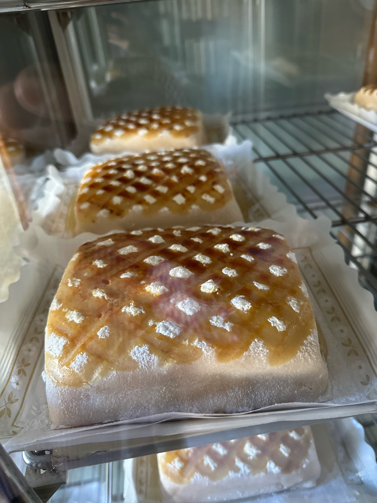 In a refrigerated display case in a bakery located in La Lastrilla, Spain, are multiple marzipan cakes. The marzipan cakes are rectangular and covered in a white powdery substance, possibly powdered sugar. The top of each cake is glazed with a golden-brown glaze that has been scored in a diamond pattern. The glaze has a slightly translucent appearance, revealing what appears to be a darker layer underneath in some areas. Small, white flecks of what could be powdered sugar or meringue are visible within the glaze, particularly along the scored lines. The cakes are arranged in rows, with at least three visible in the foreground, each on a white paper liner with a subtle gold floral pattern. The background shows the interior of the display case, including metal shelving and reflections of people and the surrounding environment. The lighting is bright, highlighting the texture and sheen of the marzipan cakes.