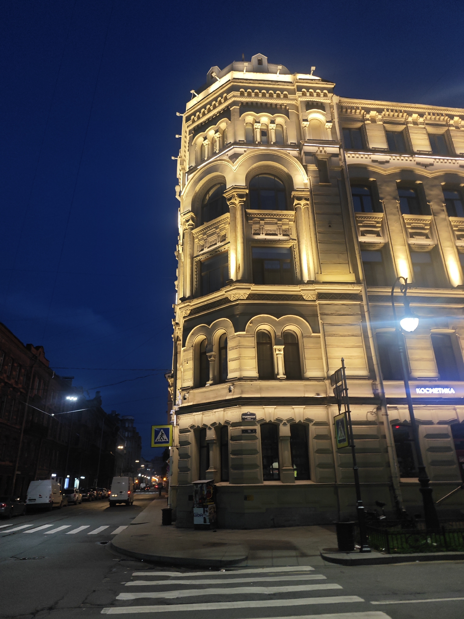 The image captures a street corner in Saint Petersburg, Russia, at twilight. A grand, multi-story building with ornate architectural details and exterior lighting dominates the right side of the frame. The building's facade is a light color, possibly stone or stucco, and is illuminated by warm yellow lights highlighting its decorative elements, including arched windows and intricate cornices. A street lamp with a glowing white bulb stands near the building.
The street in front of the building is paved and shows white pedestrian crossing stripes. On the left side of the image, another building with darker brickwork and fewer architectural embellishments lines the street. Several white vans and other vehicles are parked or driving along the street, suggesting it is an active urban area. Overhead, power lines crisscross the dark blue, twilight sky. A yellow pedestrian crossing sign with a silhouette of a person walking is visible on a pole. The overall impression is of a well-lit historic city street as night begins to fall.