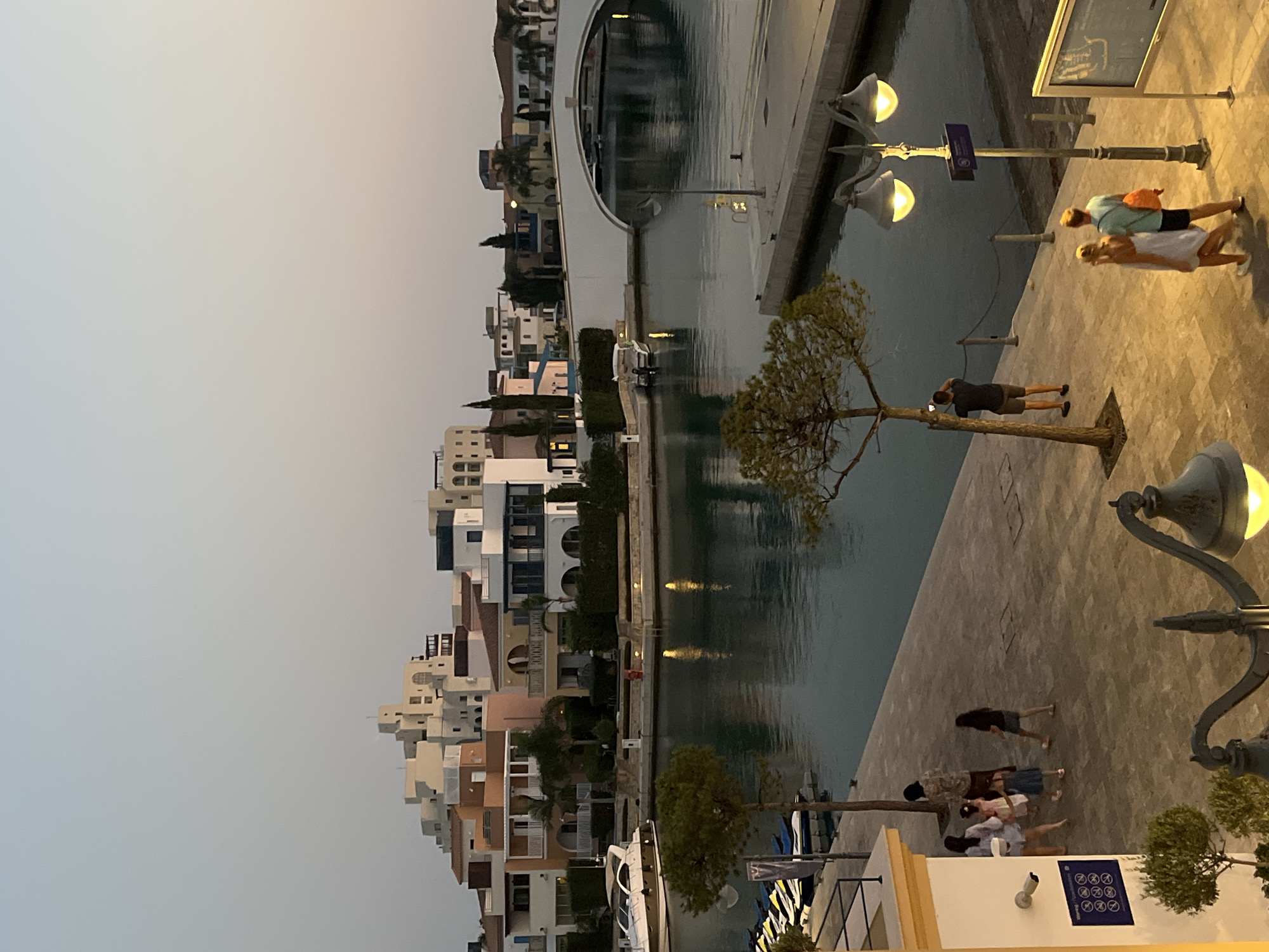 This image depicts a picturesque canal-side scene in the Limassol Marina, Cyprus, during the twilight hours. A waterway runs through the center of the frame, reflecting the soft light of the evening sky and the illuminated buildings. On the left bank, a collection of multi-story residential buildings, characterized by their modern architecture and light-colored facades, line the waterfront. A sleek white motorboat is docked on the left, and a row of jet skis is visible further along the bank.
Across the canal, a white arched bridge spans the water, connecting to another section of the marina with similar contemporary buildings. The right bank features a paved promenade where several individuals are casually strolling. A couple walks hand-in-hand in the foreground, while further back, a man stands by a tree, appearing to take a photograph with his phone. A group of three women and two children are also walking along the promenade.
Streetlights with warm yellow glows illuminate the paved walkways, casting a gentle light on the scene. The sky is a soft gradient of pale blue and peach, indicating either sunrise or sunset. The overall atmosphere is serene and inviting, showcasing the modern amenities and relaxed ambiance of the Limassol Marina.