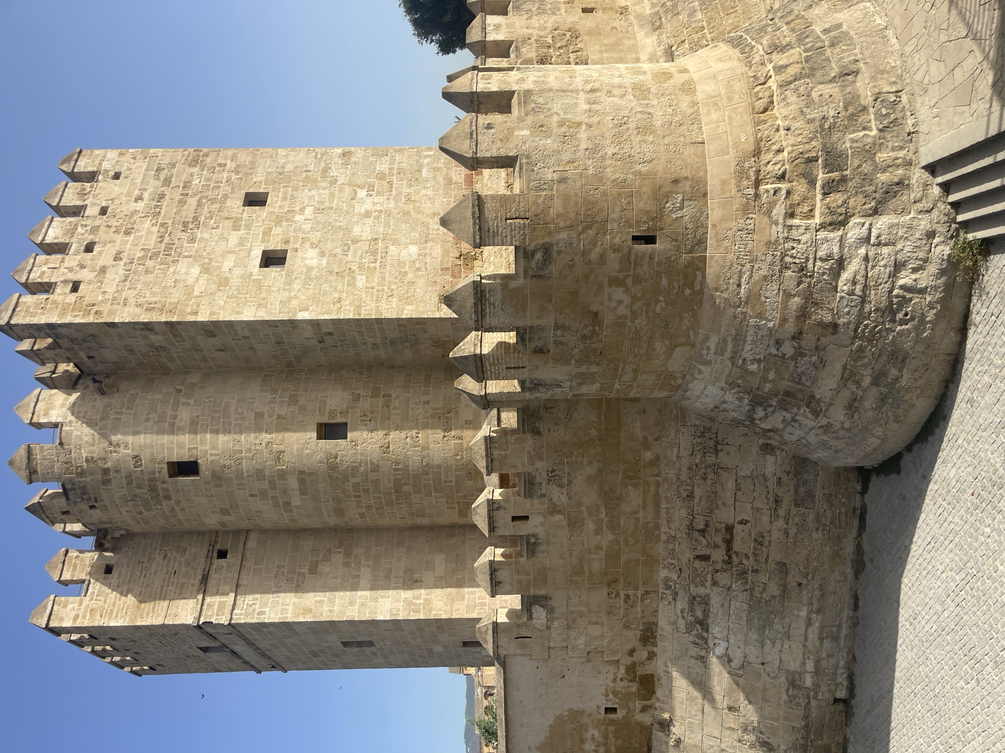 The image depicts the Torre de la Calahorra, a fortified gatehouse in Córdoba, Spain, on a clear day with a bright blue sky. The tower, constructed from light-colored stone blocks, is the dominant subject, exhibiting a crenellated top and several small, rectangular windows. A lower, circular defensive wall with similar battlements encircles the base of the tower. The ground in front of the structure is paved with cobblestones, showing a distinct shadow cast by the tower. In the background, glimpses of other stone buildings and greenery can be seen, suggesting an urban environment. Two small dark birds are visible in flight in the upper left portion of the image. The scene is devoid of any people or discernible actions.