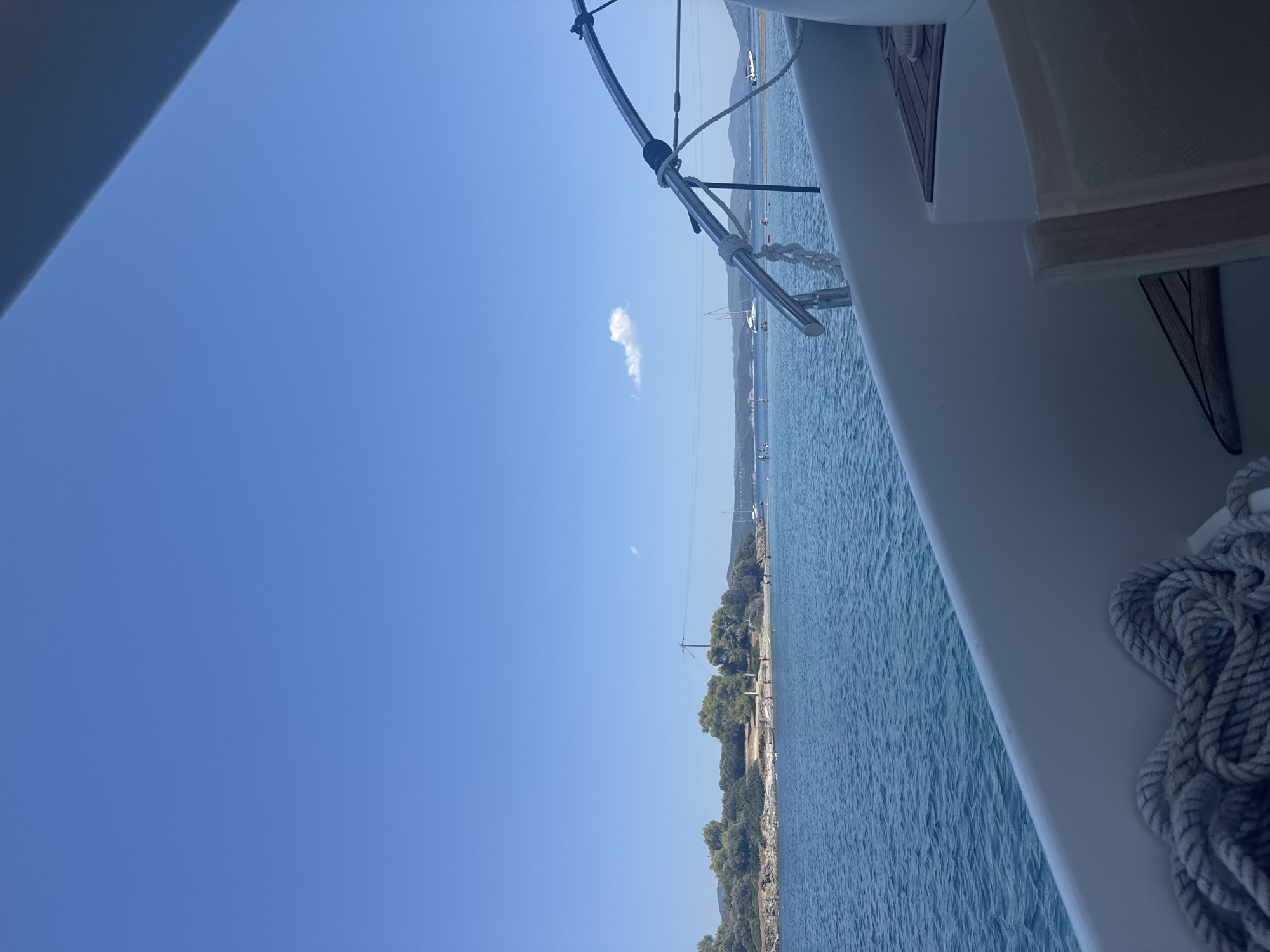 The image shows a view from a boat in the clear blue waters of Sithonia, Greece. The foreground features the deck of a white boat with a thick coiled rope. In the midground, the blue sea extends to a small, green, rocky island dotted with trees and a single utility pole. Several people are visible on the island and in the water. A few sailboats and a small motorboat are anchored in the distance. The background is dominated by a clear blue sky with a small white cloud. The overall scene depicts a serene coastal environment on a sunny day.