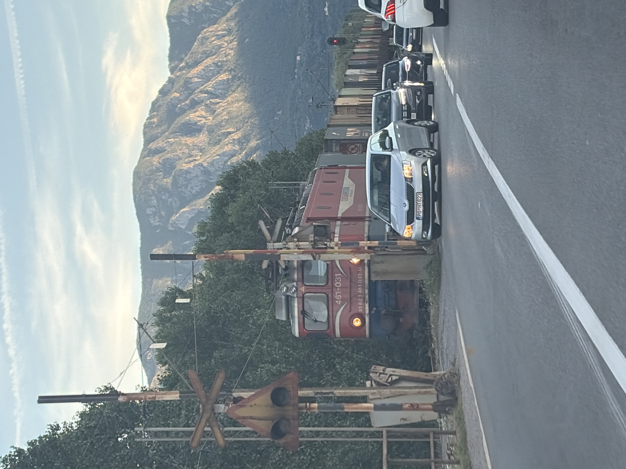 The image captures a detailed scene at a railway crossing near Virpazar, Montenegro, during either early morning or late afternoon light. In the foreground, a two-lane asphalt road runs from the bottom center towards the background, featuring a solid white line on the right and a dashed white line in the center. To the left, a weathered railroad crossing signal stands, comprising an X-shaped crossbuck sign and a triangular yellow warning sign with two circular dark lenses, mounted on vertical poles.
In the midground, a red electric locomotive, identified by the numbers "461-031" and "91 62 1 461 031-0" on its side, is partially visible behind trees and the crossing signal. The locomotive's front lights are illuminated, and it is pulling a long line of diverse freight wagons extending into the background. A queue of vehicles is stopped on the road, facing away from the viewer. The lead car is a silver Skoda Fabia with the Montenegrin license plate "PG HT623" (indicating Podgorica registration), its headlights and turn signals illuminated. A driver is visible inside. Behind it, a dark grey Volkswagen Polo is stopped, followed by other vehicles, including a dark car and the front portion of a white car on the far right. Further down the road, a red traffic light is visible for the road traffic. Dense green foliage, consisting of trees and bushes, lines the railway tracks and the immediate roadside.
The background is dominated by large, rugged mountains, displaying exposed rock formations and sparse vegetation, characteristic of the Montenegrin landscape. The sky above is a light blue with scattered, thin white clouds, including faint contrails. The overall lighting is soft, with hints of golden light on the mountain peaks, suggesting either sunrise or sunset.