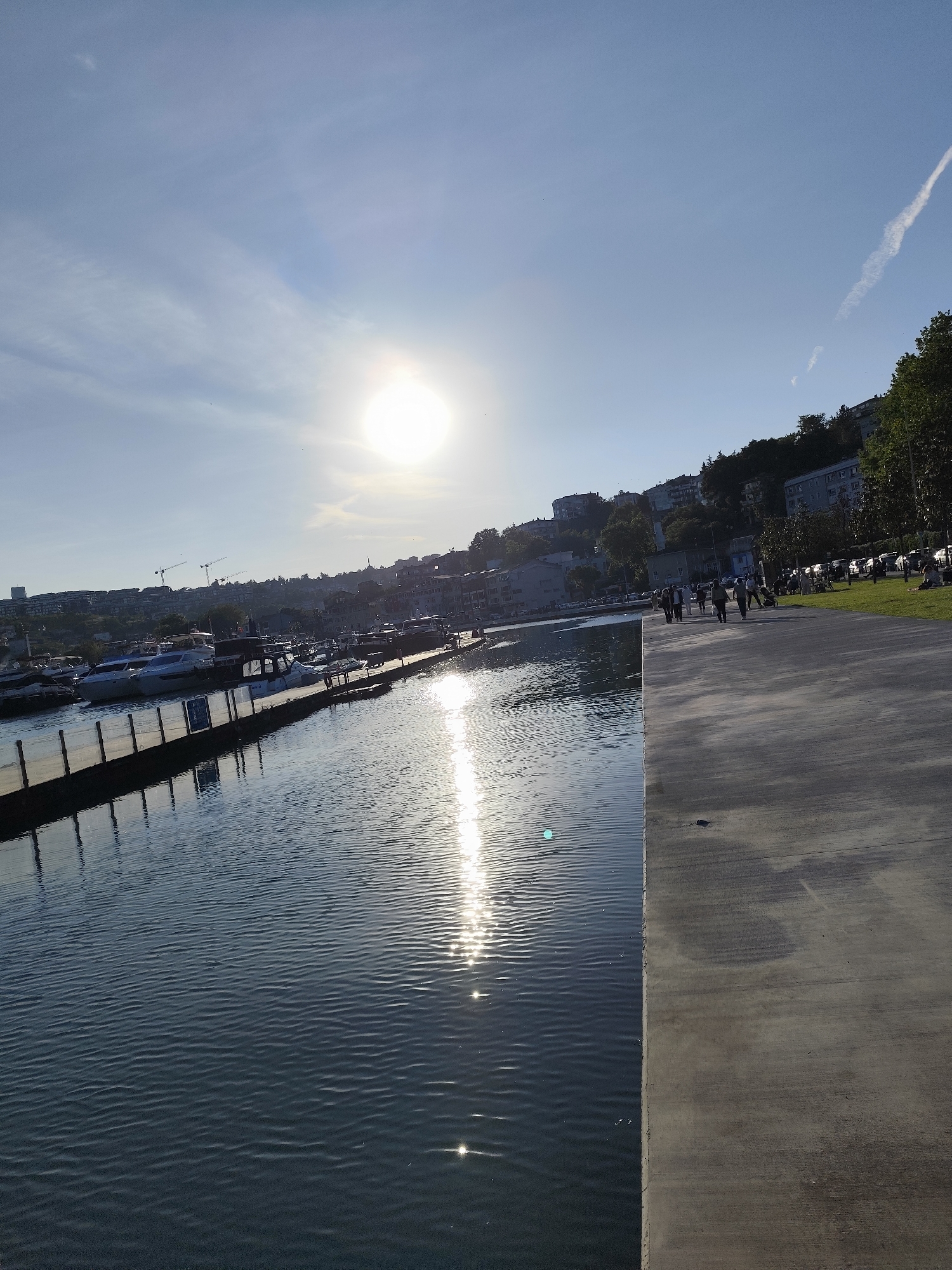 The image depicts a sunny scene in Sarıyer, Turkey, likely along the coastline. The sun is high in the sky, causing glare on the water's surface. There is a harbor with moored boats. A concrete walkway runs along the edge of the water. Some people are walking on the walkway. The background shows a hillside with buildings and trees.