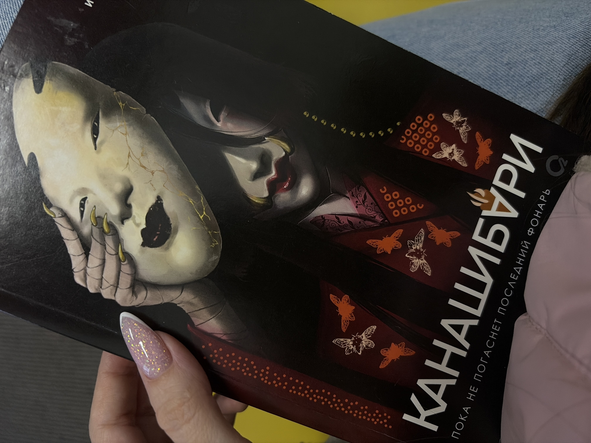 A person is holding a book titled "Канашибури" (Kanashiburi) which translates to "while the light does not extinguish the flashlight," written in Russian. The book cover features a woman holding a mask with long, sharp nails. The person holding the book has a glittery nail design with a white tip. The scene appears to be indoors. The location, Saint Petersburg, Russia, cannot be determined from the image.